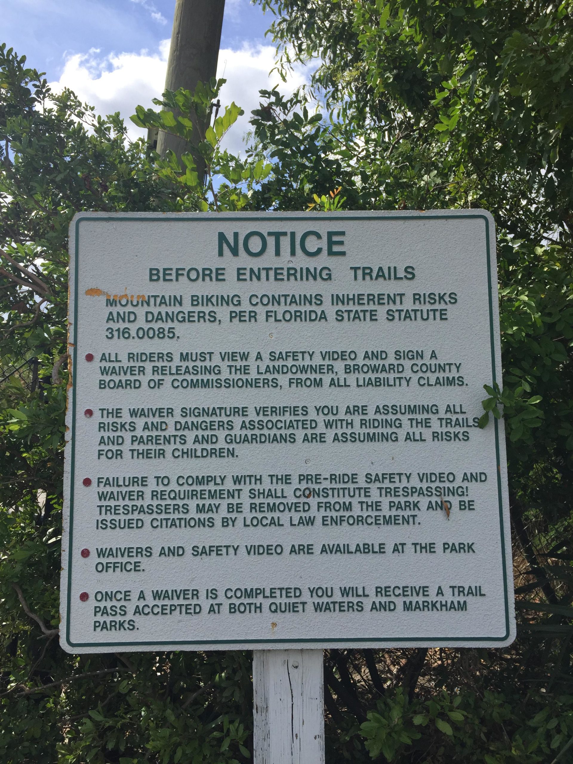 A sign posted near mountain biking trails, detailing safety precautions and legal disclaimers. The notice warns that mountain biking carries inherent risks and requires riders to view a safety video and sign a waiver that releases liability for the landowner and the Broward County Board of Commissioners. It also states that failure to comply may result in removal from the park, and outlines the process for obtaining a trail pass. The sign is surrounded by greenery and has a blue sky in the background. Quiet Waters Park mountain bike trail.