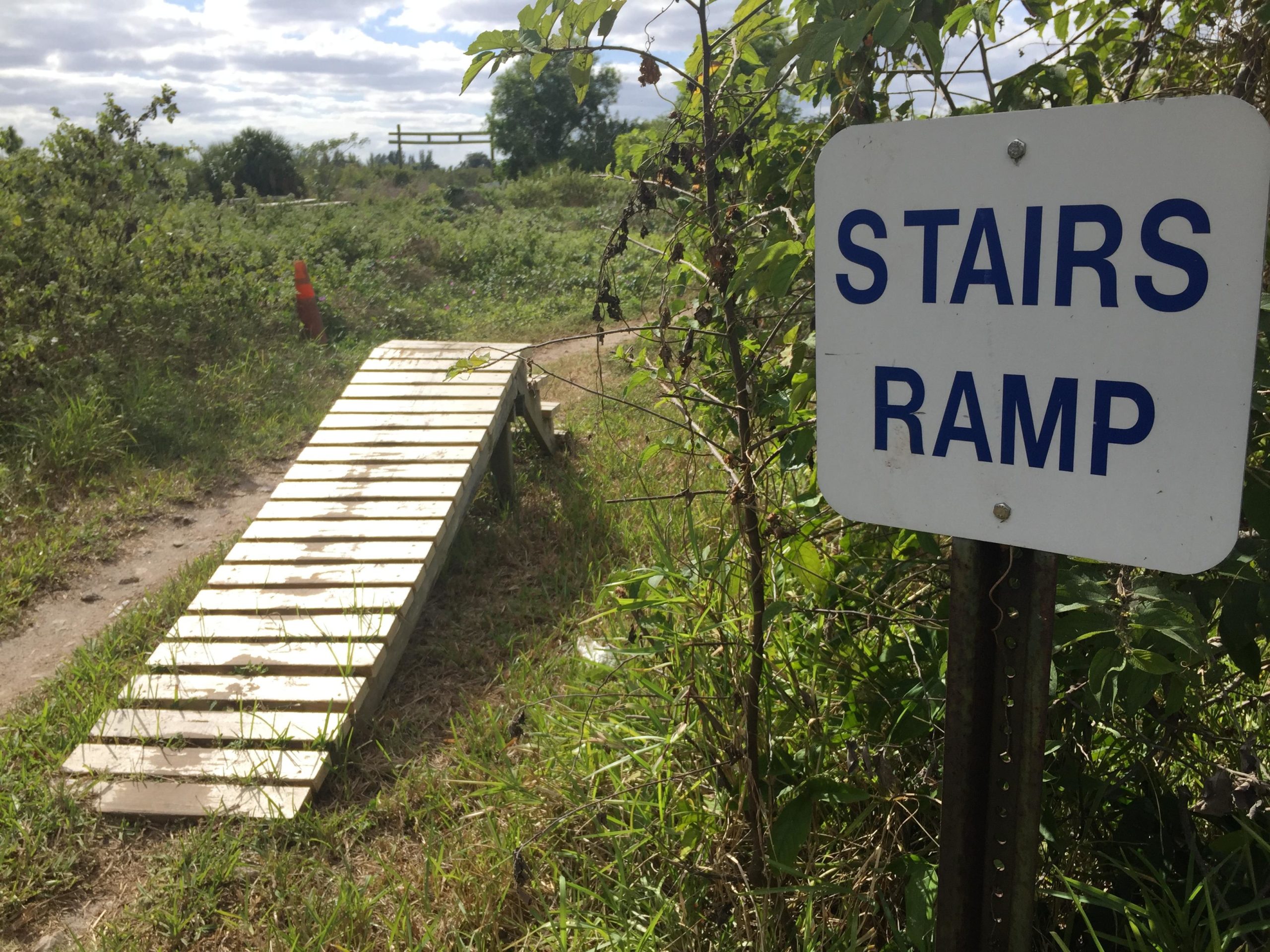A wooden ramp leading from a dirt path into a grassy area, next to a sign that reads "STAIRS RAMP." The background features green foliage and a cloudy sky. A traffic cone is positioned nearby. Quiet Waters Park mountain bike trail.
