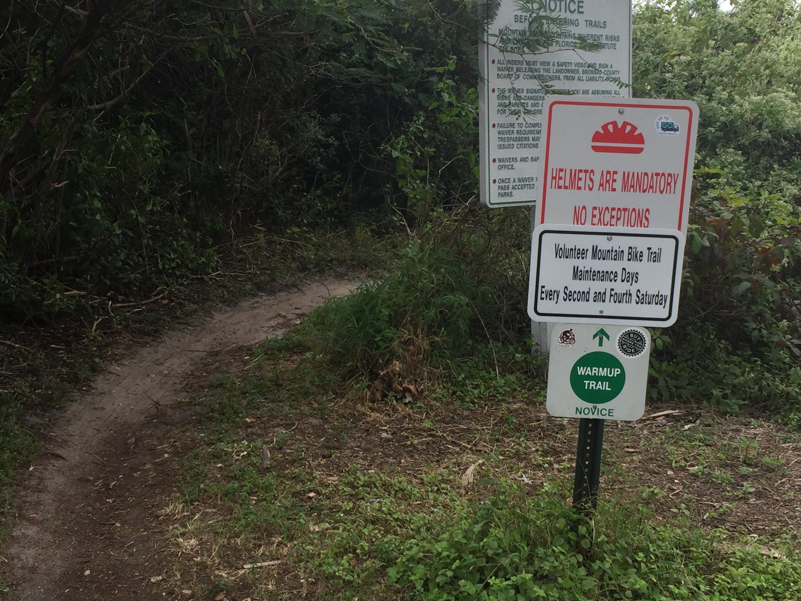 A winding dirt path leads into a wooded area, accompanied by multiple informational signs. One sign emphasizes that helmets are mandatory for riders, while another indicates it's part of a volunteer mountain bike trail. There are details about maintenance days occurring on the second and fourth Saturday of each month. A third sign marks the beginning of the "Warmup Trail," categorized as novice. Lush greenery surrounds the trail, creating a natural setting. Quiet Waters Park mountain bike trail.