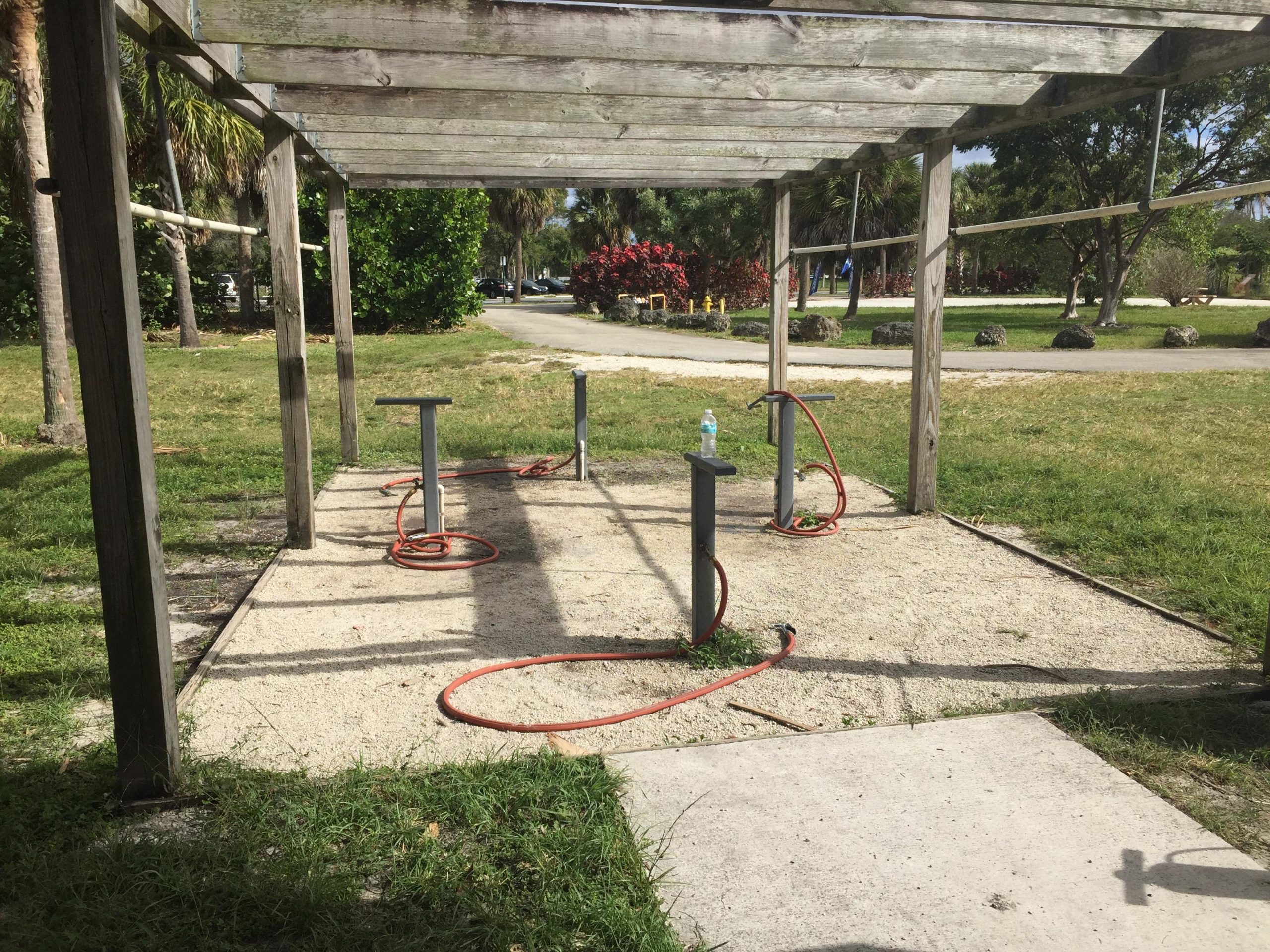 A shaded pavilion with wooden beams overlooks a sandy area equipped with hoses and posts, surrounded by green grass and palm trees. In the background, pathways and flower beds are visible. Quiet Waters Park mountain bike trail.