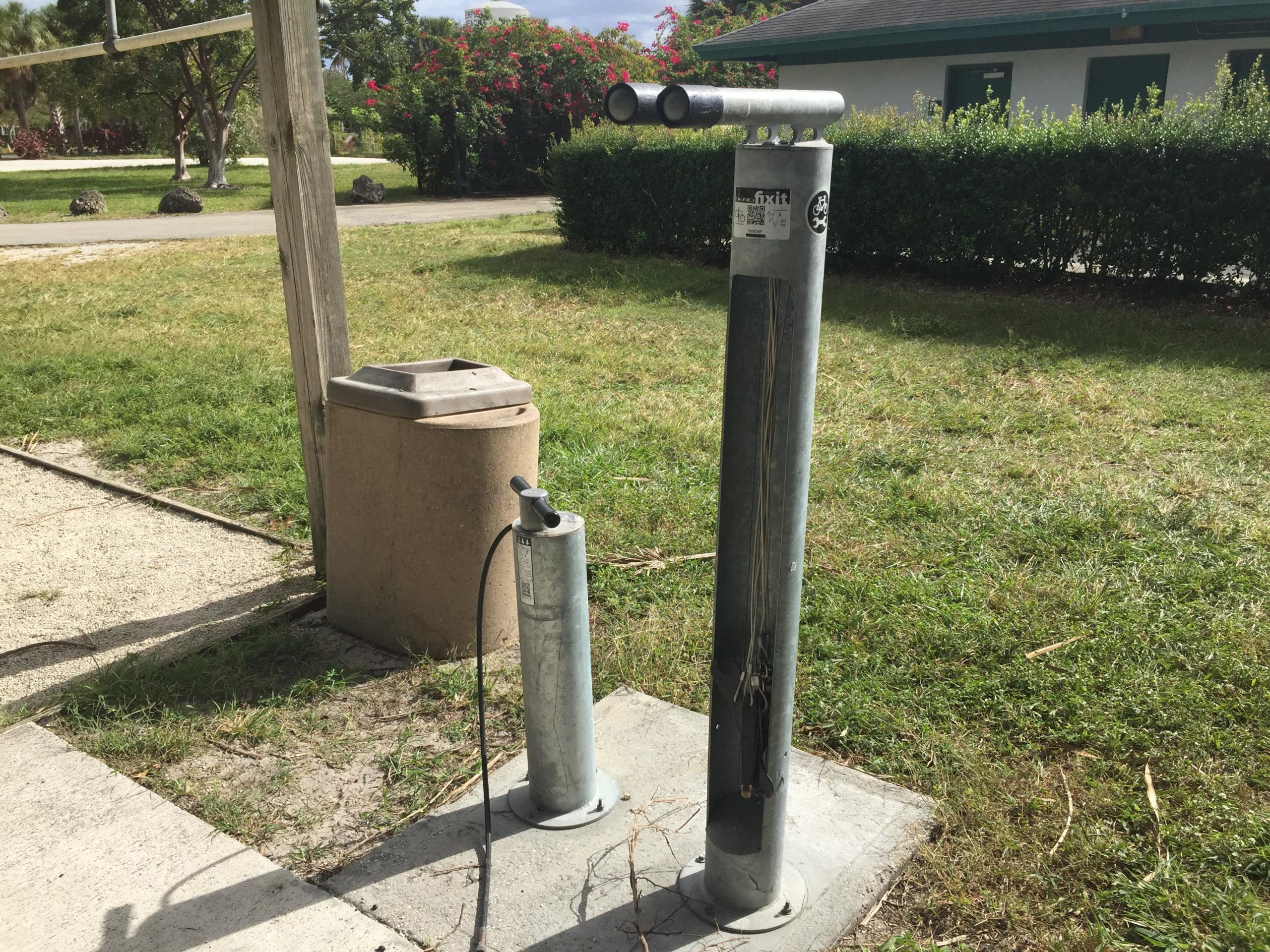 A bike repair station featuring a bicycle pump and tool holder, situated near a trash bin on a grassy area. In the background, there are trees and a building with closed doors. Quiet Waters Park mountain bike trail.