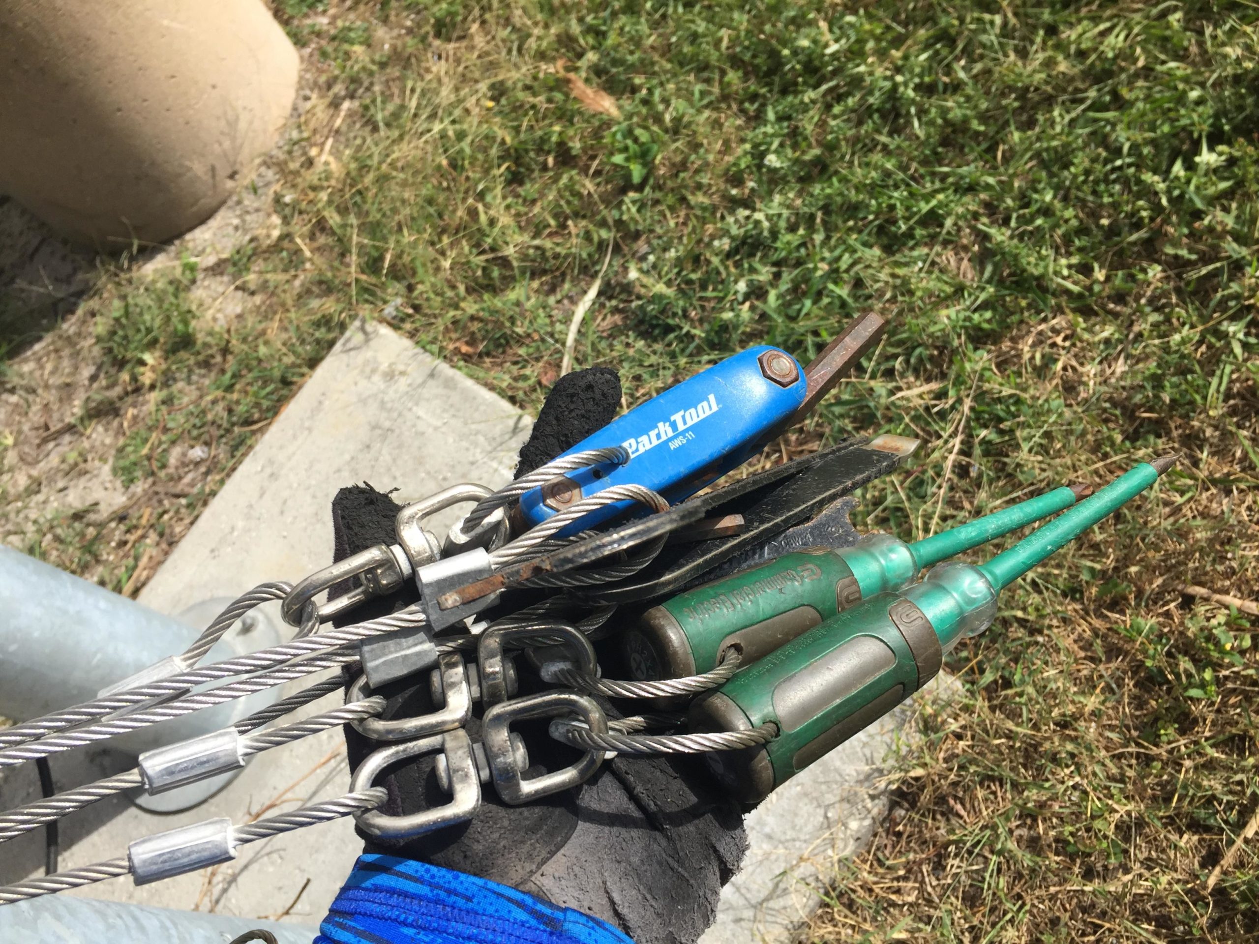 A close-up image of a gloved hand holding various tools, including a blue Park Tool multi-tool and two green screwdrivers, alongside metal cables and connectors. The background features grass and a concrete surface. Quiet Waters Park mountain bike trail.