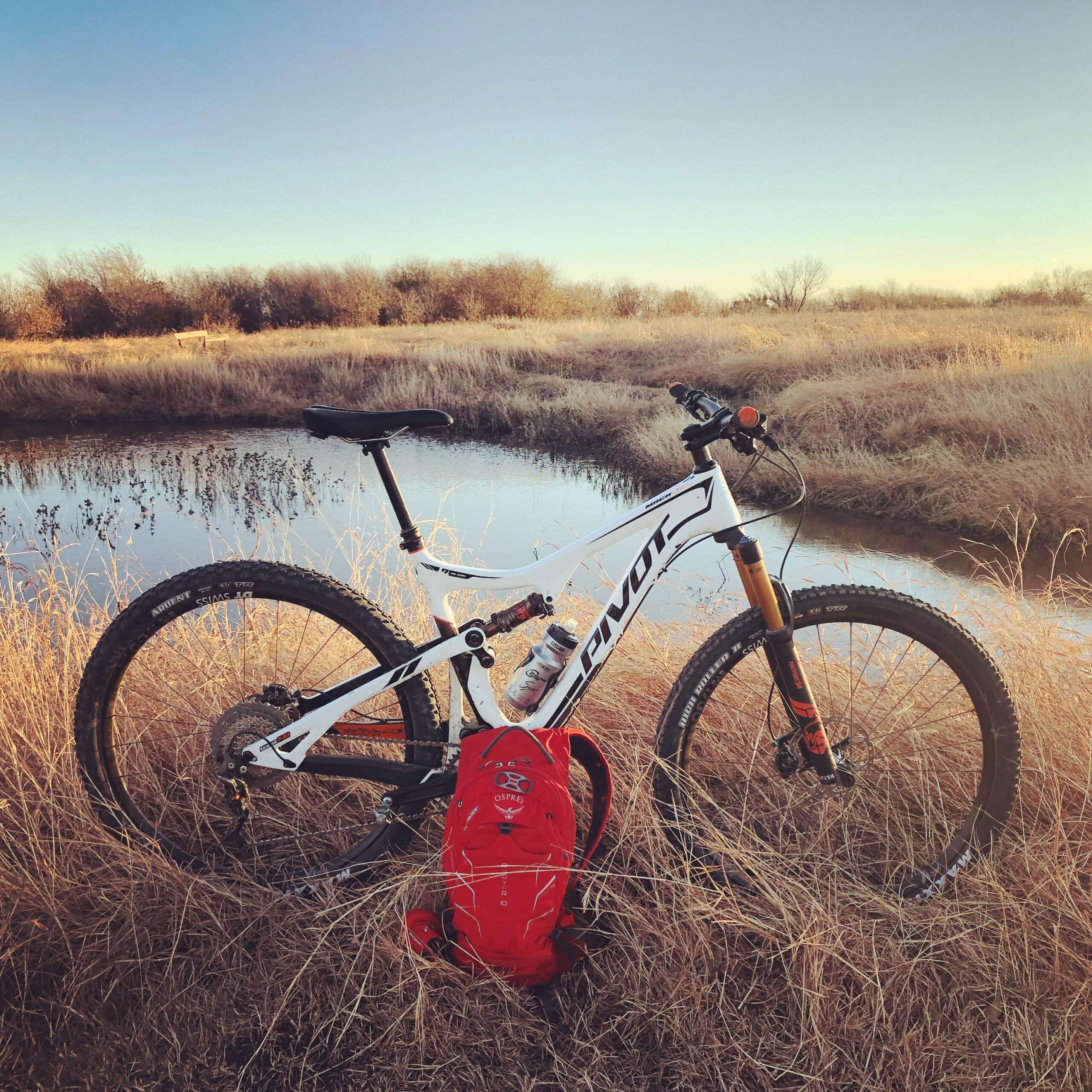 Pivot 429 Trail XT/XTR Pro: A white mountain bike leaning against a grassy area near a small pond, with a red backpack on the ground beside it. The scene is set in an open landscape with sparse trees in the background and a clear sky. The sun is setting, casting a warm glow over the surroundings.