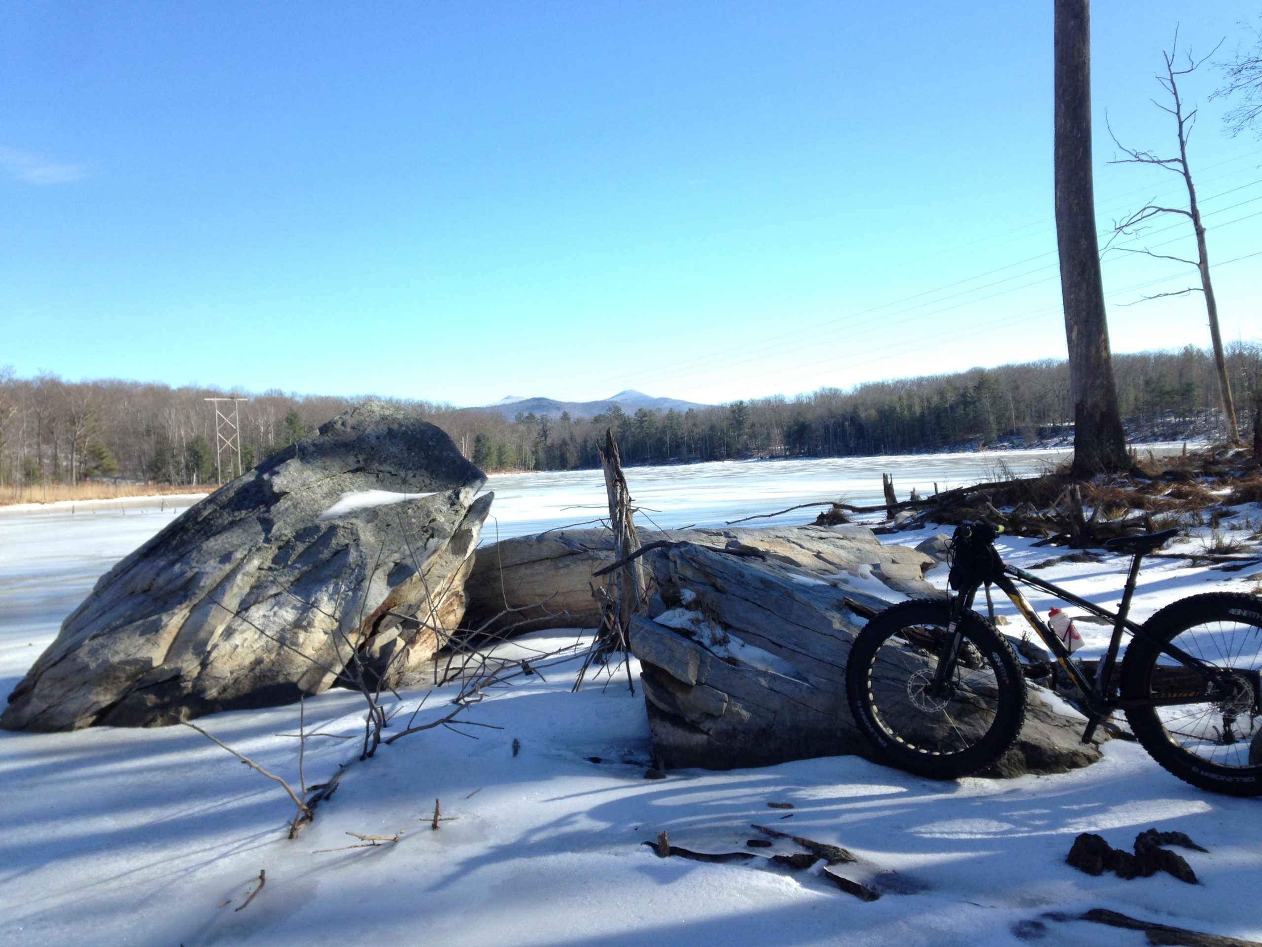 A scenic winter landscape featuring large rocks alongside a frozen lake, with a clear blue sky overhead. In the foreground, a mountain bike rests on the snow-covered ground. Beyond the lake, there are trees and distant mountains, creating a serene outdoor setting. Pine Hill Park mountain bike trail.
