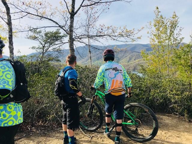 Two mountain bikers stand on a trail overlooking a scenic view of mountains and a body of water. One rider is looking at the view while the other is checking the bike. Both are wearing colorful biking jerseys and gear, surrounded by trees and greenery. Tsali Recreation Area mountain bike trail.