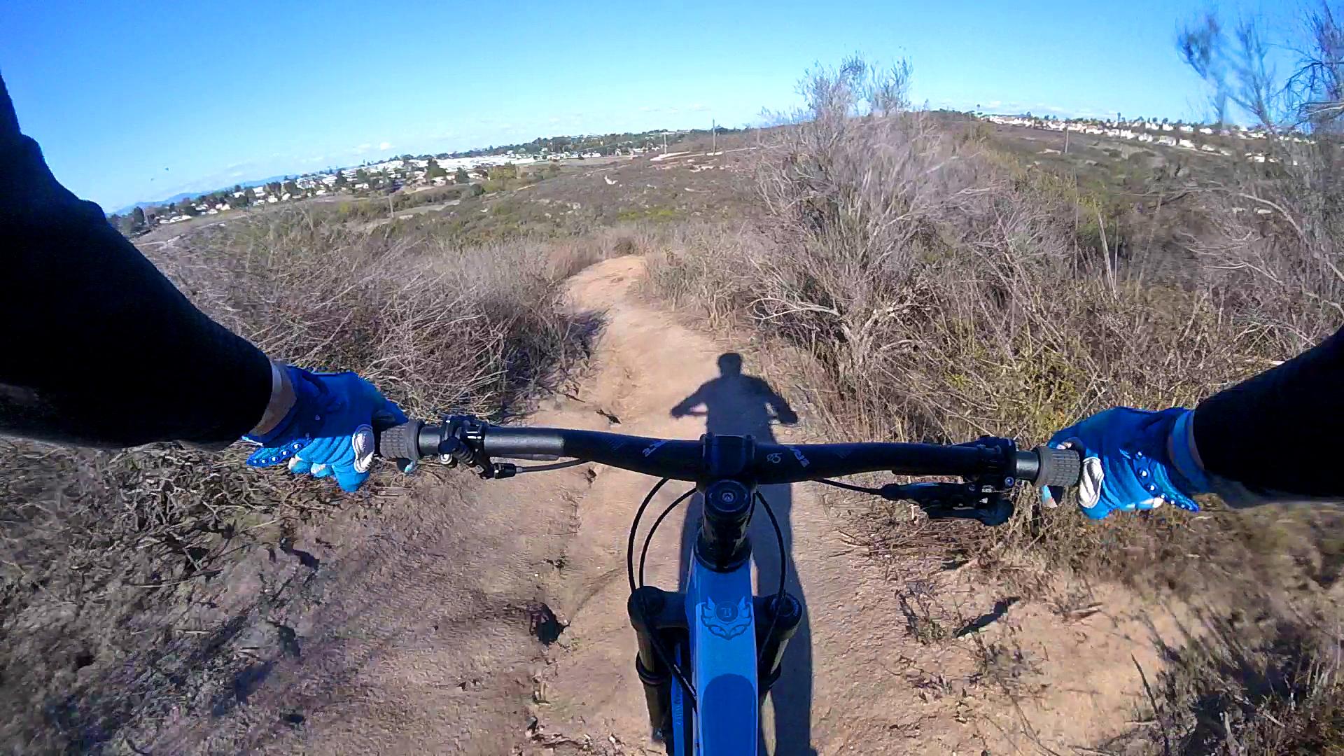A view from the handlebars of a mountain bike riding along a dirt trail, with a clear blue sky above and a distant view of houses and landscape. The cyclist's shadow is visible on the ground, and the bike has blue accents. Surrounding the trail are sparse bushes and vegetation. Lake Calavera mountain bike trail.