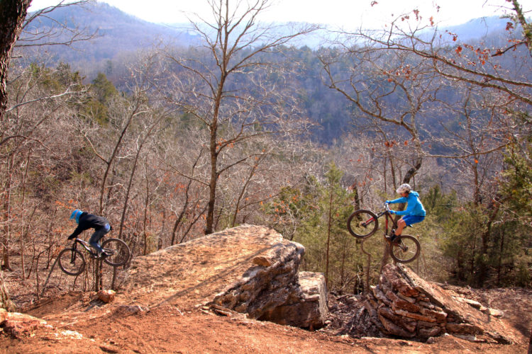 Two mountain bikers are performing tricks on rocky terrain in a forested area. One rider is jumping over a large stone while the other is maneuvering on a rock formation. The background features bare trees and a mountain landscape under a clear sky, indicating a crisp outdoor setting.