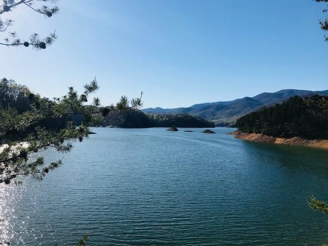 A tranquil lake surrounded by mountains and trees under a clear blue sky. The water reflects the natural scenery, with small islands visible in the distance. Tsali Recreation Area mountain bike trail.