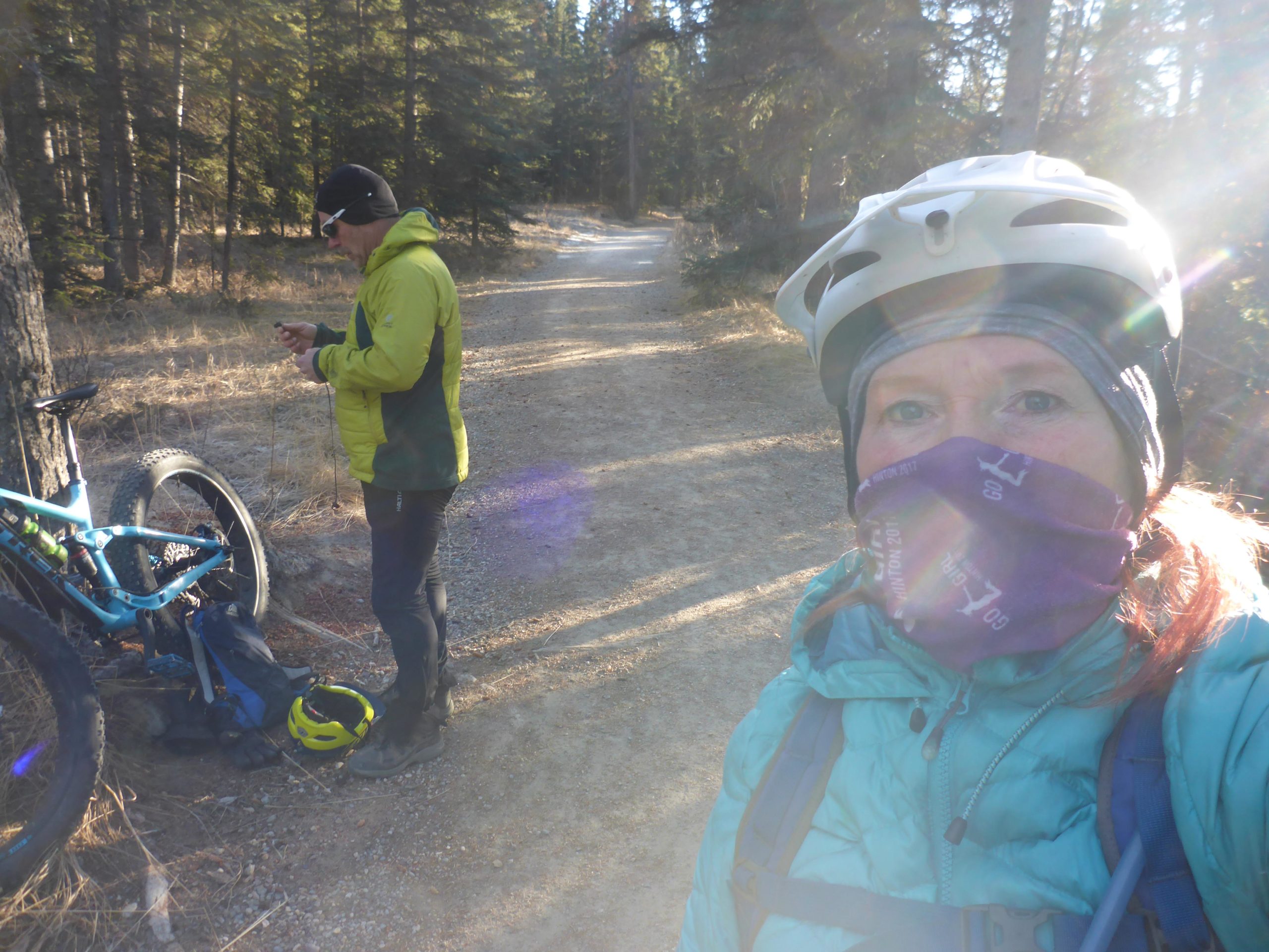 A woman wearing a helmet and a blue jacket takes a selfie while standing on a gravel path in a forest. In the background, a man in a green jacket is next to a mountain bike and appears to be checking his phone. The scene is illuminated by sunlight filtering through the trees. Trail 7 / 7H (Teahouse) mountain bike trail.