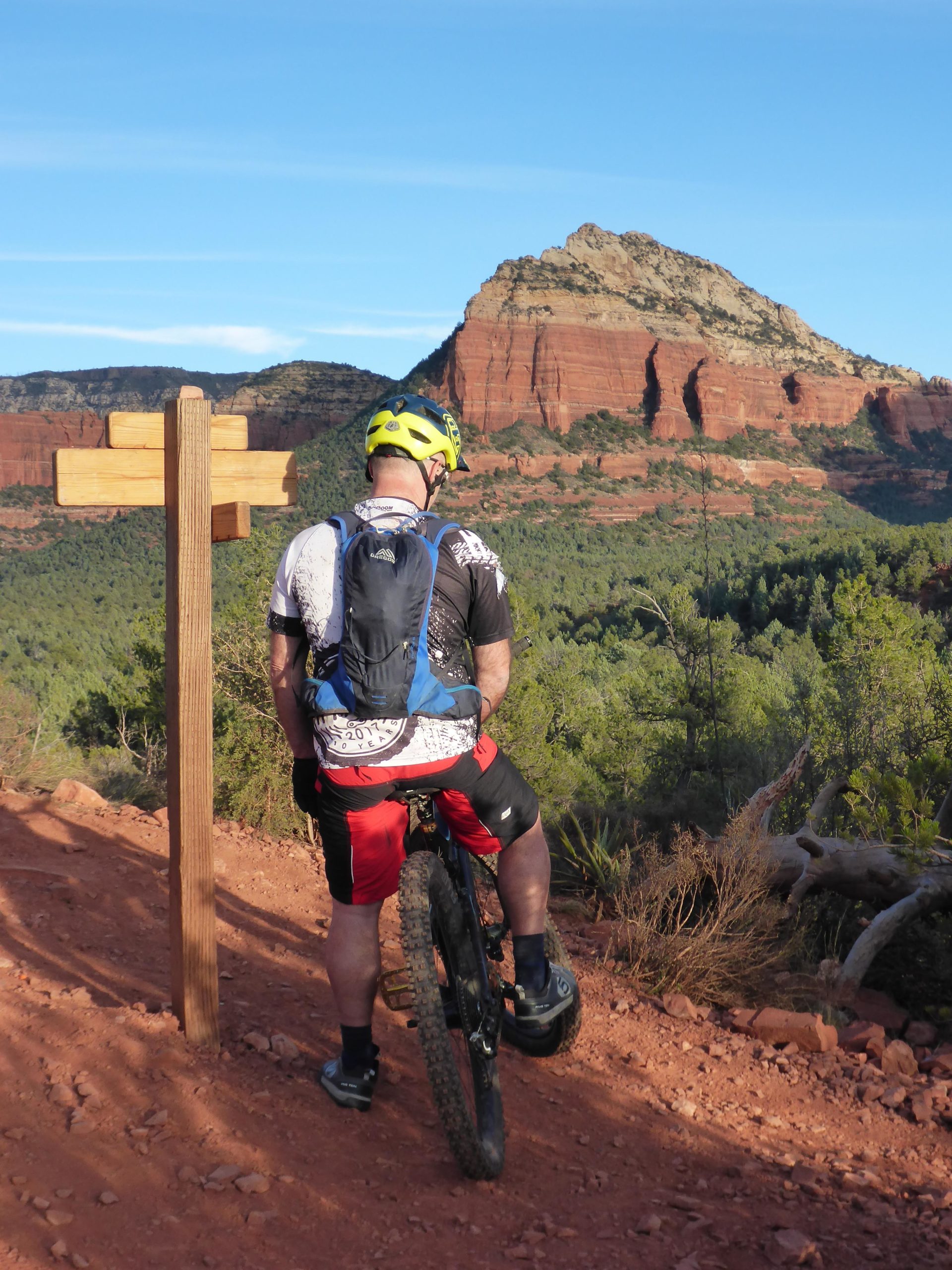 A mountain biker in a yellow helmet and a black and white cycling jersey stands beside a wooden trail sign, overlooking a scenic red rock landscape under a clear blue sky. Upper Dry Creek Area Trails mountain bike trail.