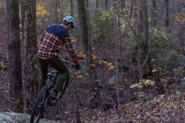 A mountain biker navigating rocky terrain in a wooded area during autumn. The rider is wearing a plaid shirt and a helmet, with trees displaying fall foliage in the background.