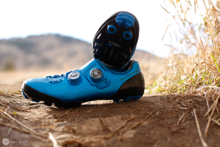 A blue Shimano cycling shoe positioned on a dirt path, with a blurred natural background. The shoe features a sleek design with two adjustment dials and a perforated upper for ventilation.