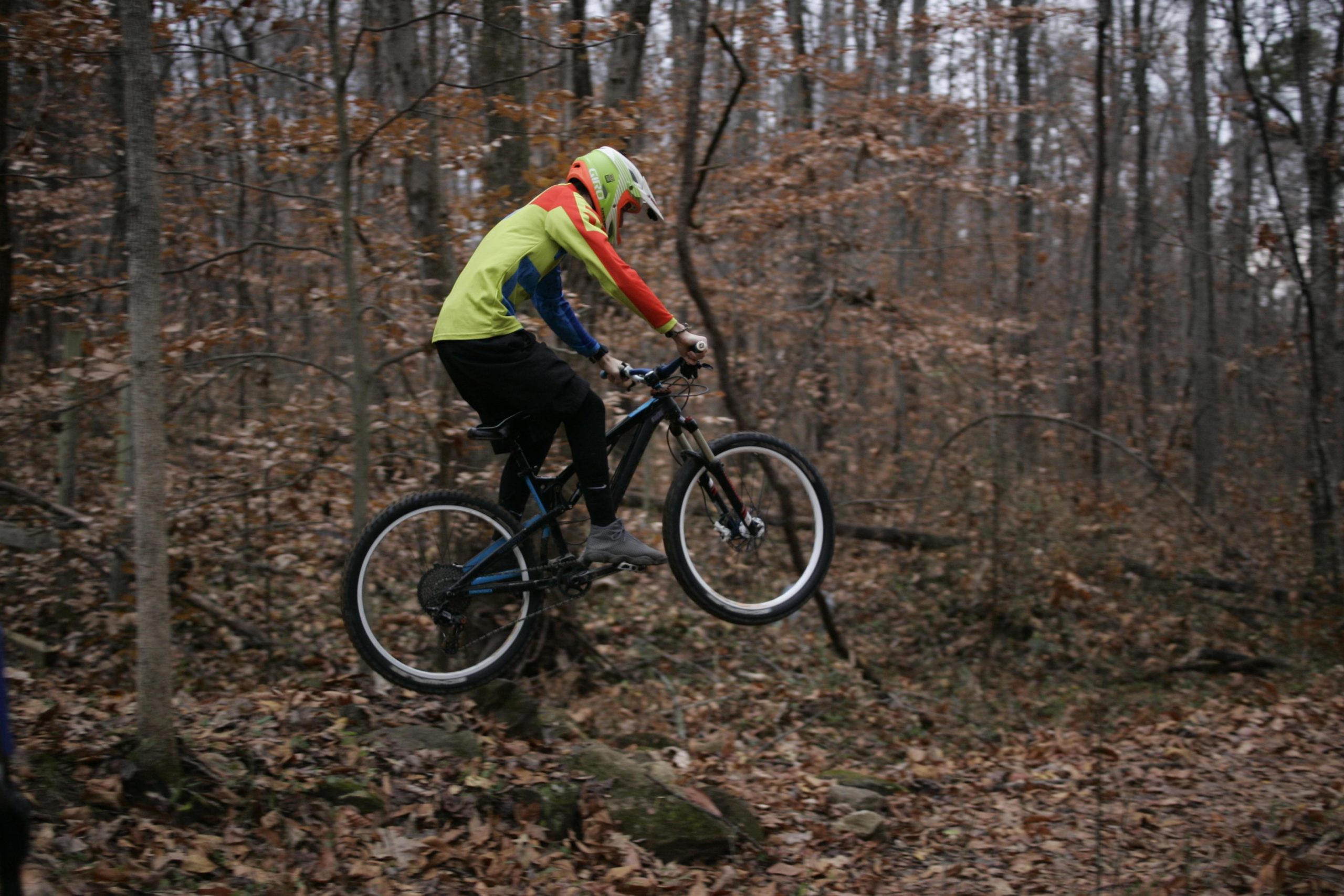 A mountain biker wearing a helmet and bright, colorful clothing jumps off a rocky terrain in a wooded area during autumn. The ground is covered with fallen leaves, and the surrounding trees have sparse foliage. The biker's bike is partially airborne as they perform the jump. Hobby Park mountain bike trail.