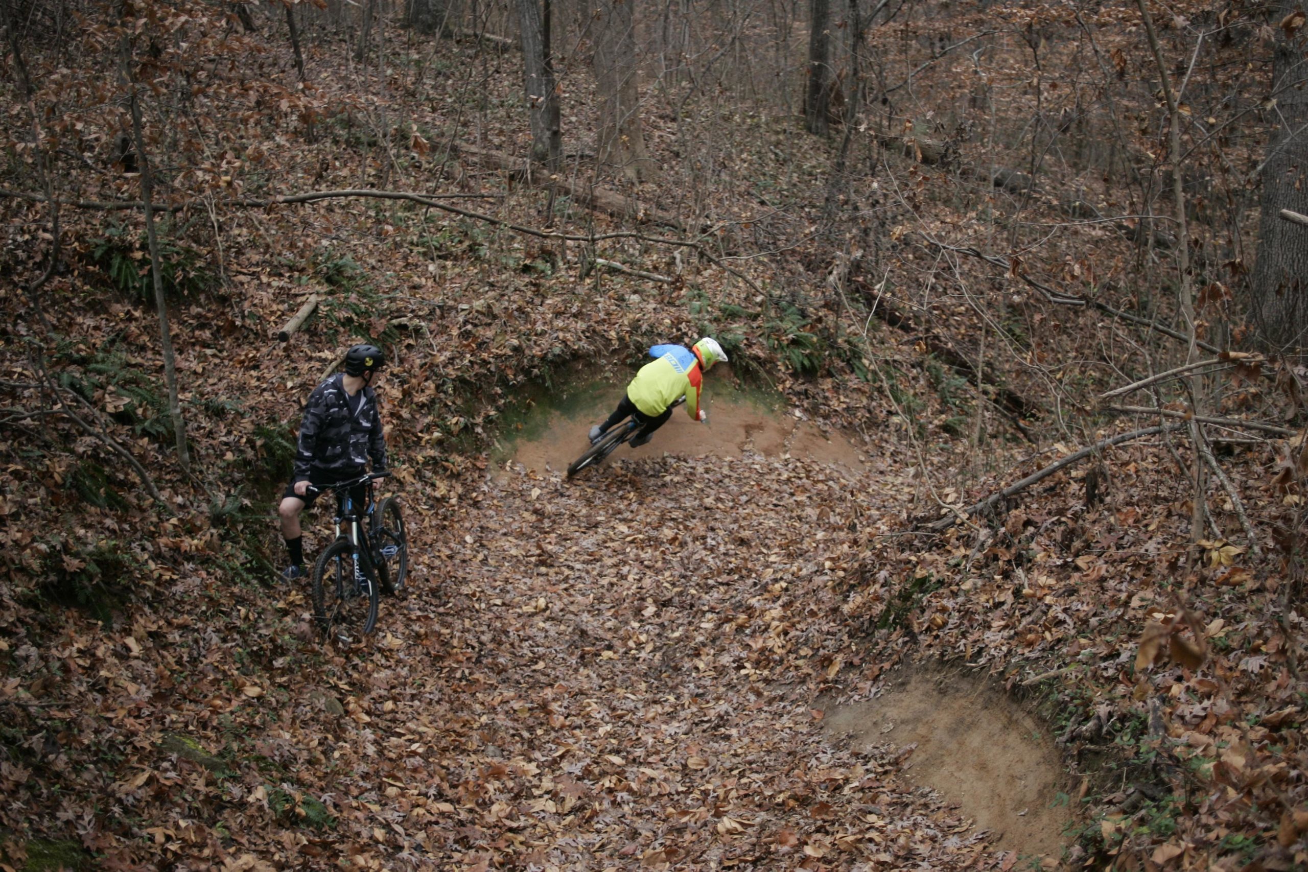 Two mountain bikers are riding in a wooded area covered with autumn leaves. One biker, dressed in a bright yellow and blue outfit, is leaning into a turn on a dirt path, while the other biker, wearing a camouflage jacket and black helmet, is standing next to his bike, observing the trail. The scene captures the essence of outdoor adventure during the fall season. Hobby Park mountain bike trail.