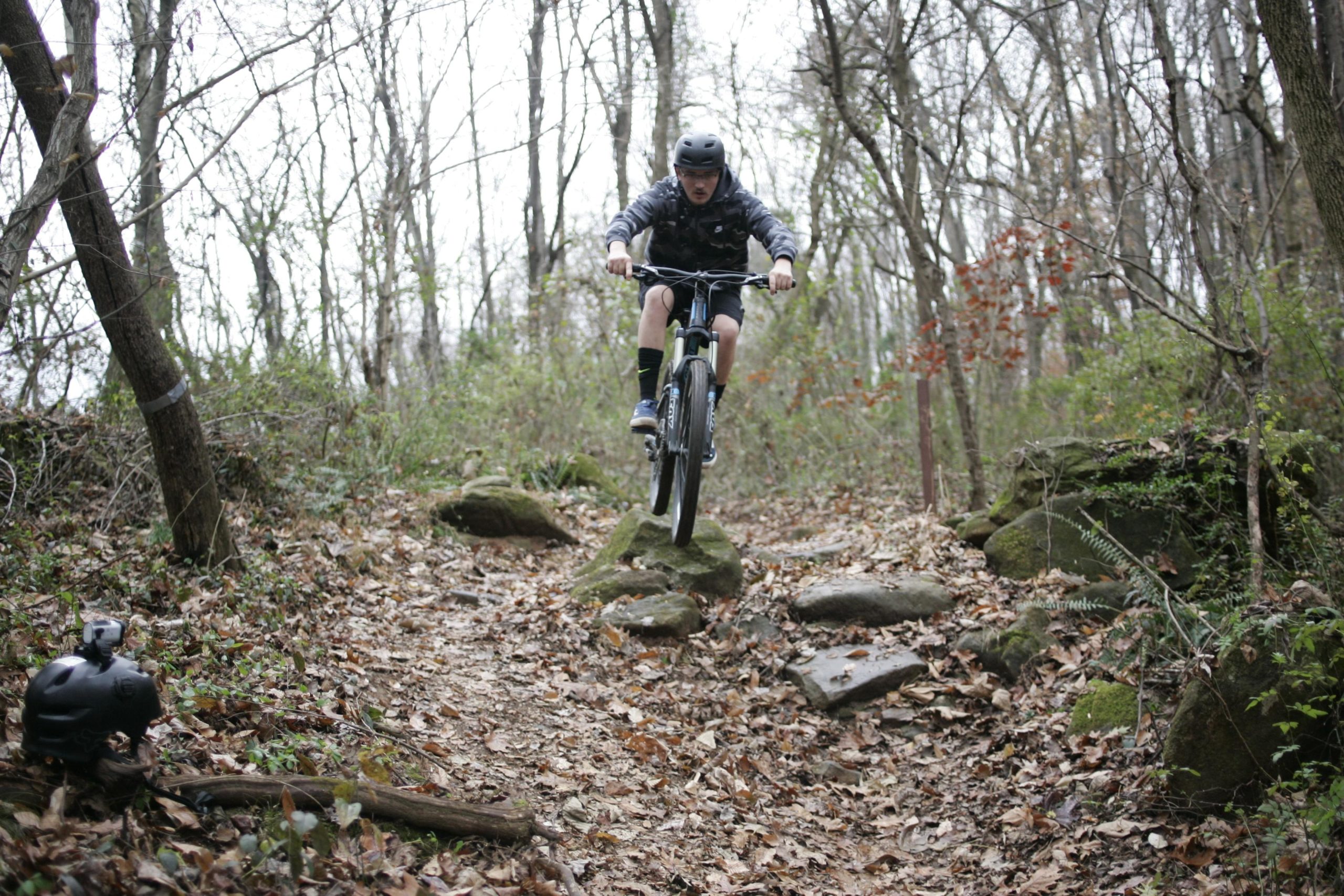 A cyclist wearing a helmet jumps off a rocky ledge on a mountain bike trail surrounded by trees and fallen leaves. A black helmet with a camera is placed on the ground nearby. Hobby Park mountain bike trail.