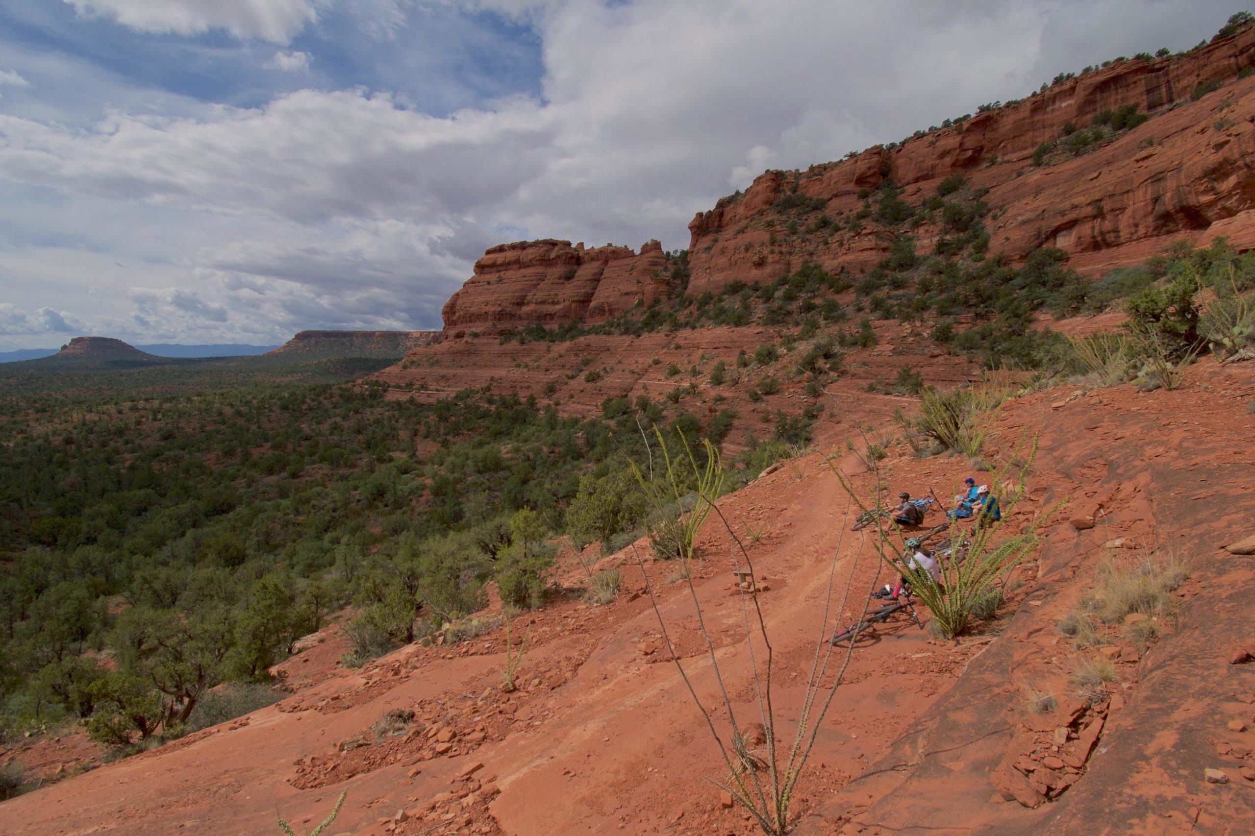 A scenic view of red rock formations and a rugged landscape, featuring two mountain bikers resting on a slope. The foreground shows desert vegetation, while the background highlights a vast expanse of hills and a cloudy sky. Mescal Trail mountain bike trail.