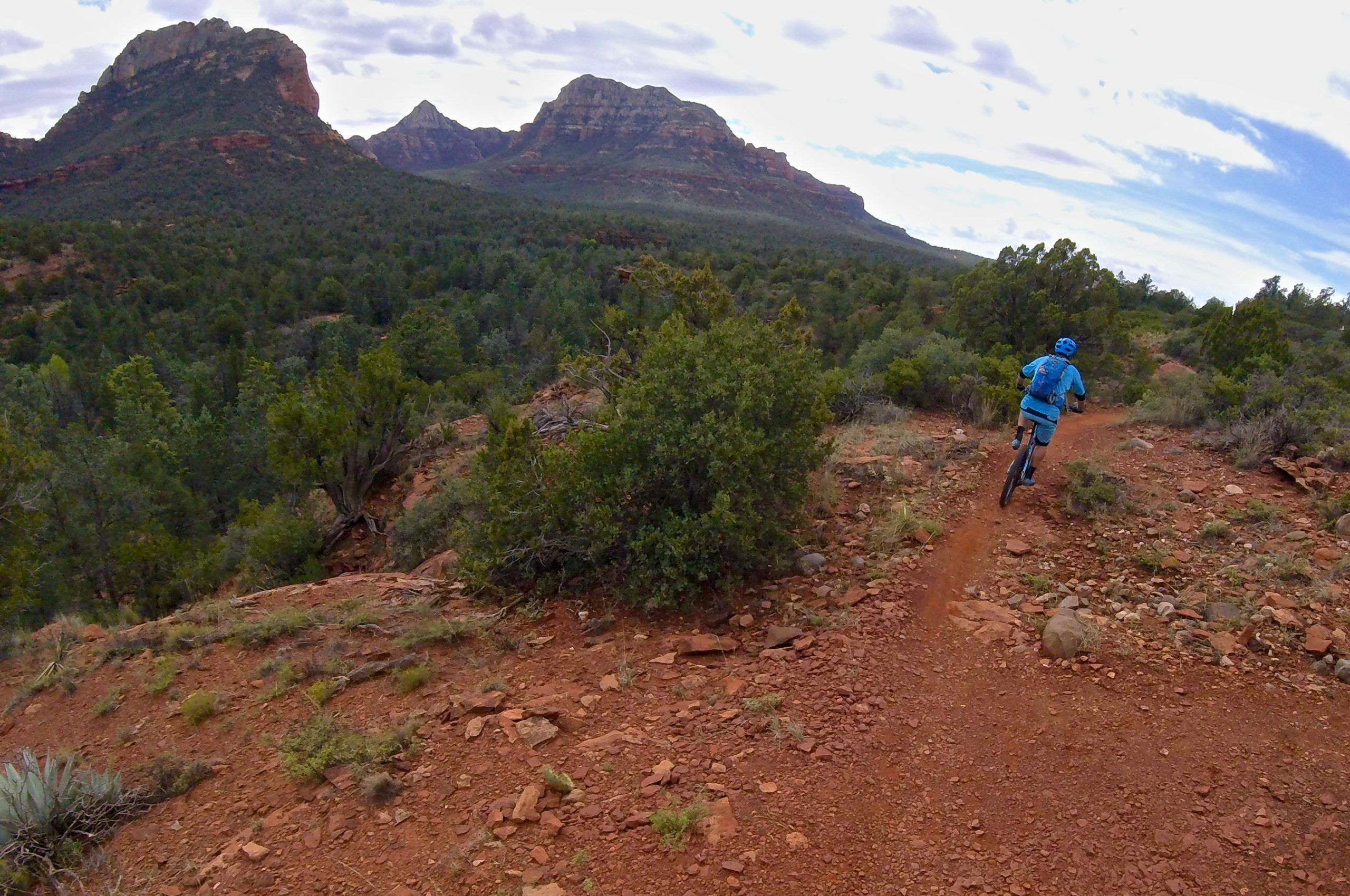 A person riding a mountain bike along a dirt trail in a rugged landscape, surrounded by green trees and rocky formations. The sky is partly cloudy, and distant mountainous peaks are visible in the background. Chuck Wagon mountain bike trail.