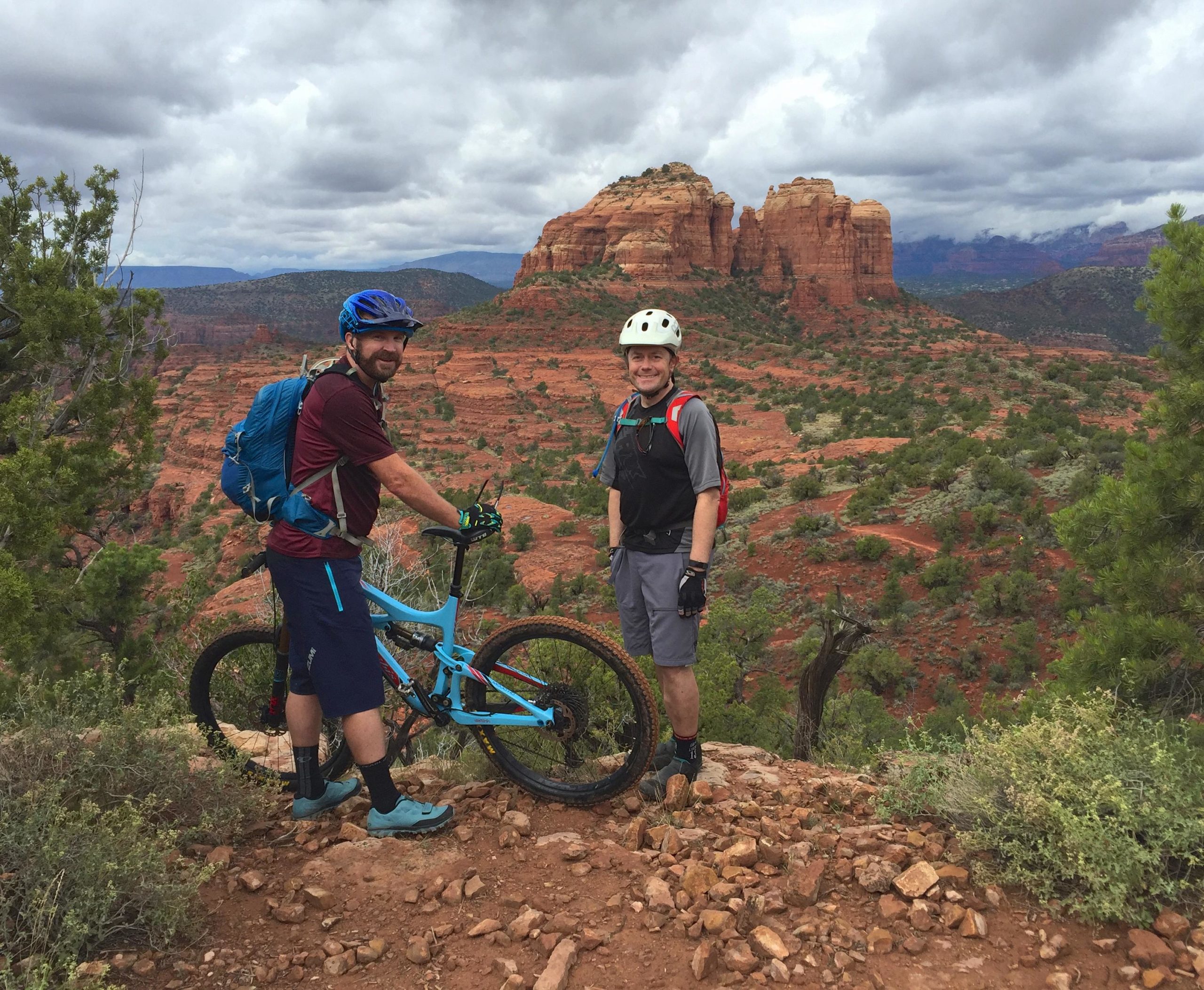 Two mountain bikers stand on a rocky overlook with a stunning view of red rock formations in the background. One rider, wearing a blue helmet and a backpack, stands next to a blue mountain bike, while the other, in a white helmet and black gear, smiles at the camera. The landscape features green shrubs and a cloudy sky above. Hiline mountain bike trail.