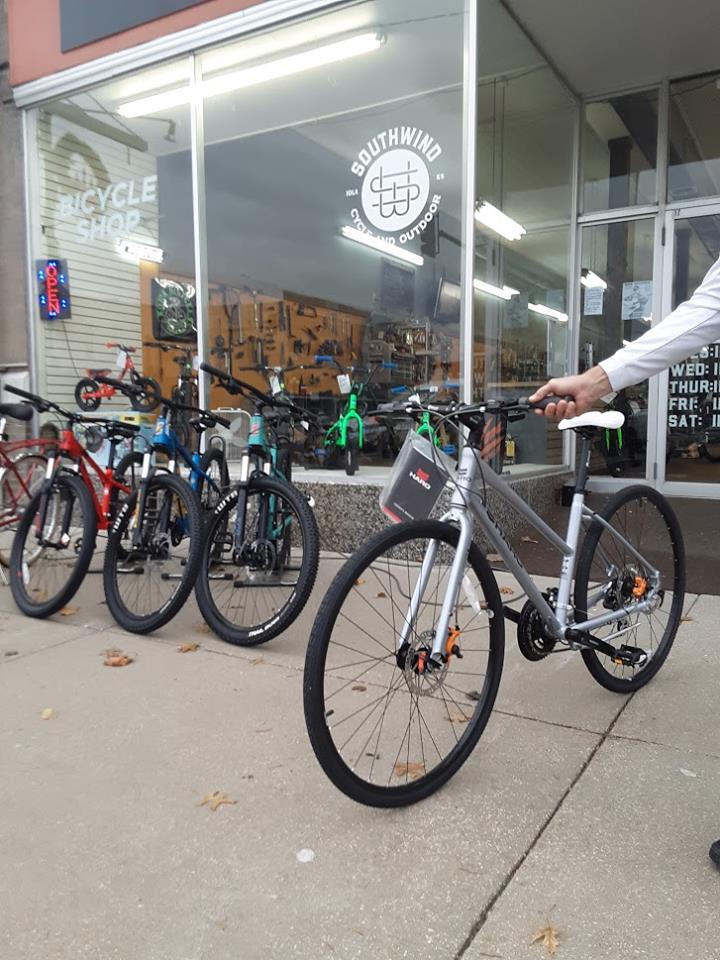 A bicycle shop storefront featuring a selection of bicycles outside. The shop, named "Southwind," displays various bikes, including a silver mountain bike in the foreground. The window shows additional merchandise and tools, with bright lighting and a colorful "Open" sign visible.