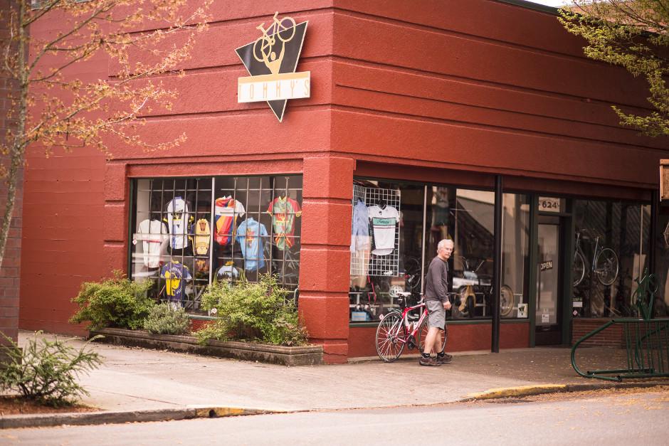 A cyclist stands outside a bike shop with a red exterior, featuring a large logo with a bicycle symbol. The store