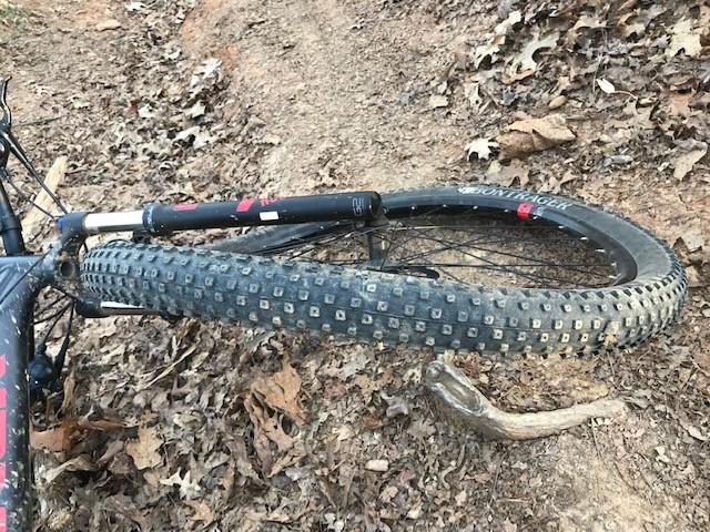 Close-up view of a bent mountain bike wheel on a dirt trail surrounded by fallen leaves. The tire features a rugged tread pattern, typical of off-road biking, and is partially resting on a small stick. The bike frame is partially visible, indicating it has been laid down or is at rest. Tsali Recreation Area mountain bike trail.