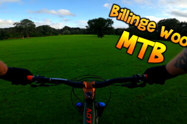 A cyclist's perspective with hands on the handlebars of a mountain bike, overlooking a grassy field surrounded by trees under a partly cloudy sky. The image features the text "Billinge Woods MTB" prominently displayed. Billinge Woods mountain bike trail.