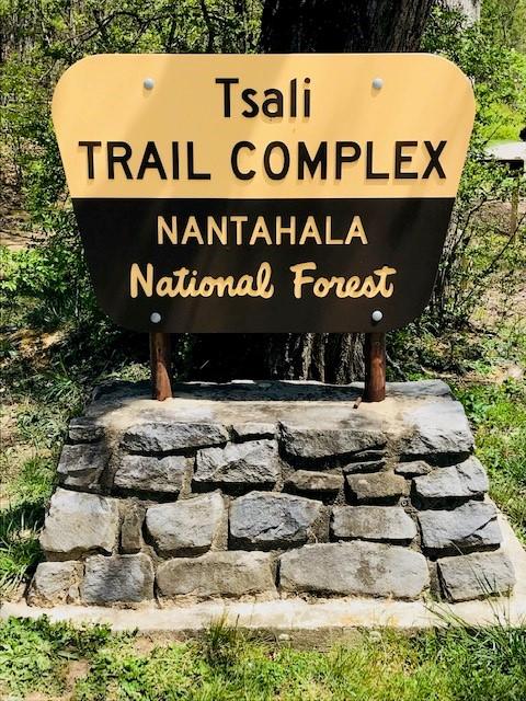 Sign for Tsali Trail Complex in Nantahala National Forest, featuring a yellow and brown design mounted on a stone base. The sign indicates the location of the trail complex within the forest. Surrounding greenery is visible. Tsali Recreation Area mountain bike trail.