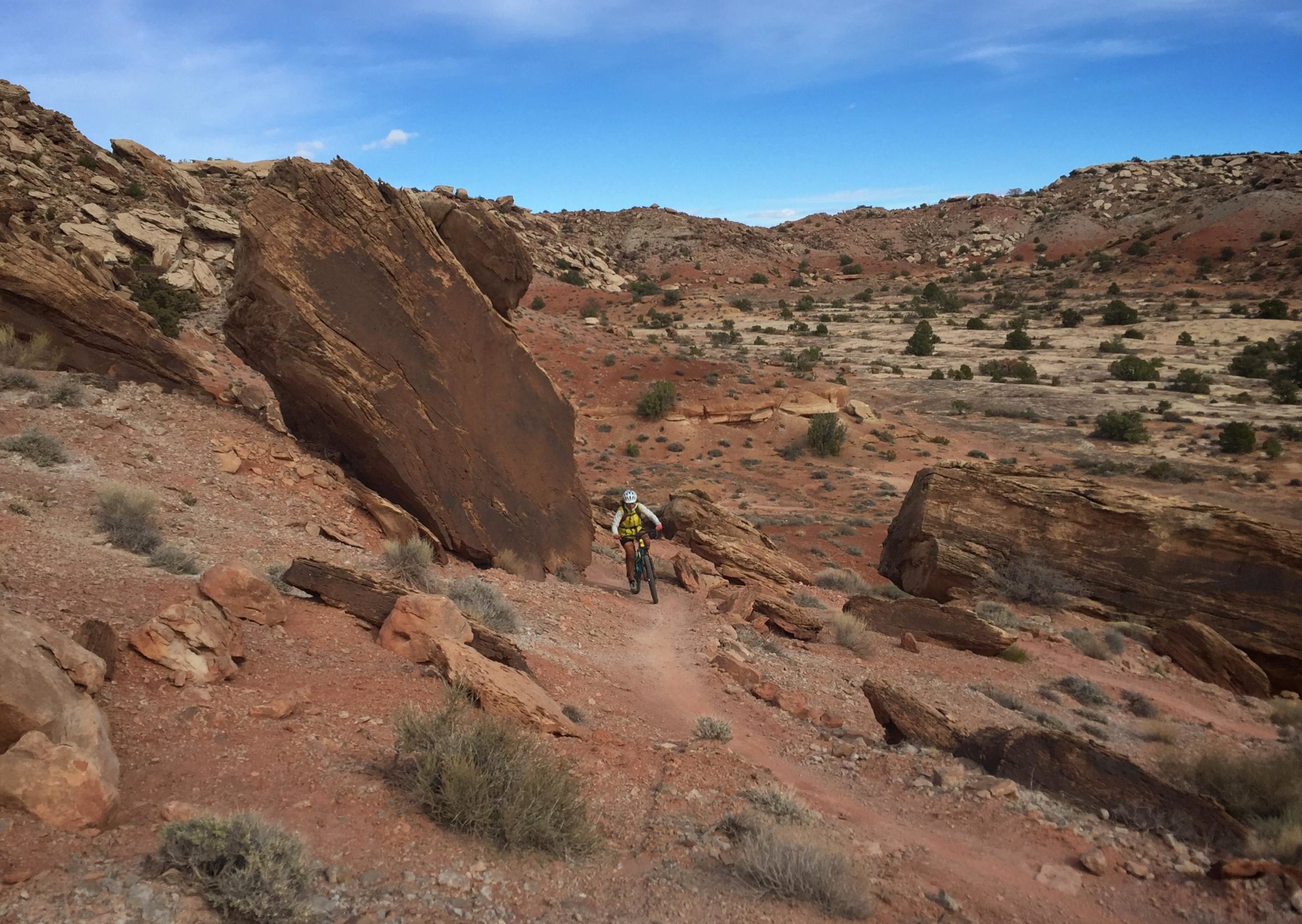 A mountain biker navigates a rocky trail in a desert landscape, surrounded by large boulders and sparse vegetation under a blue sky with scattered clouds. Klondike Bluffs mountain bike trail.