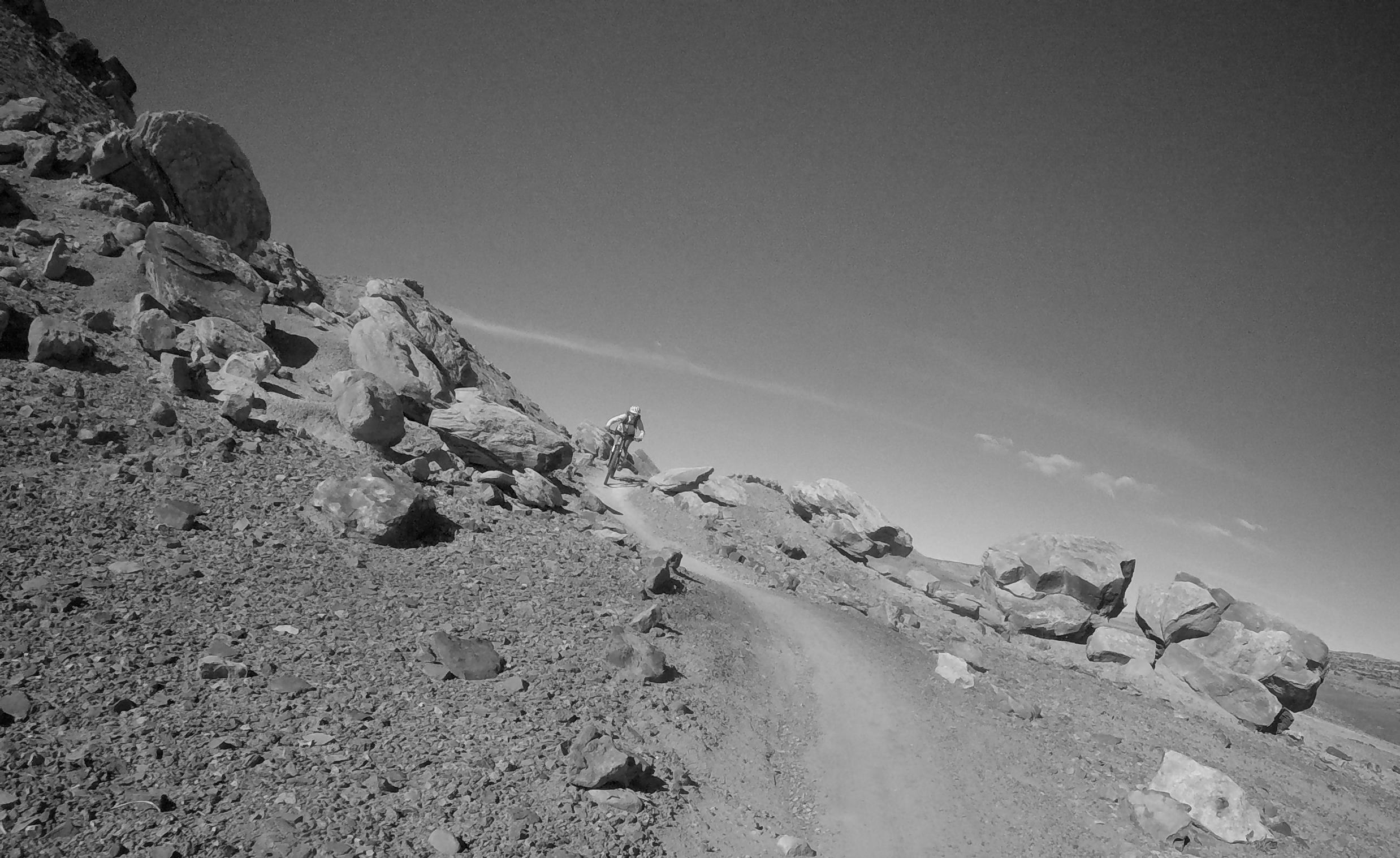 A black and white image of a mountain biker riding on a narrow dirt trail surrounded by large rocks and rugged terrain under a clear sky. Klondike Bluffs mountain bike trail.