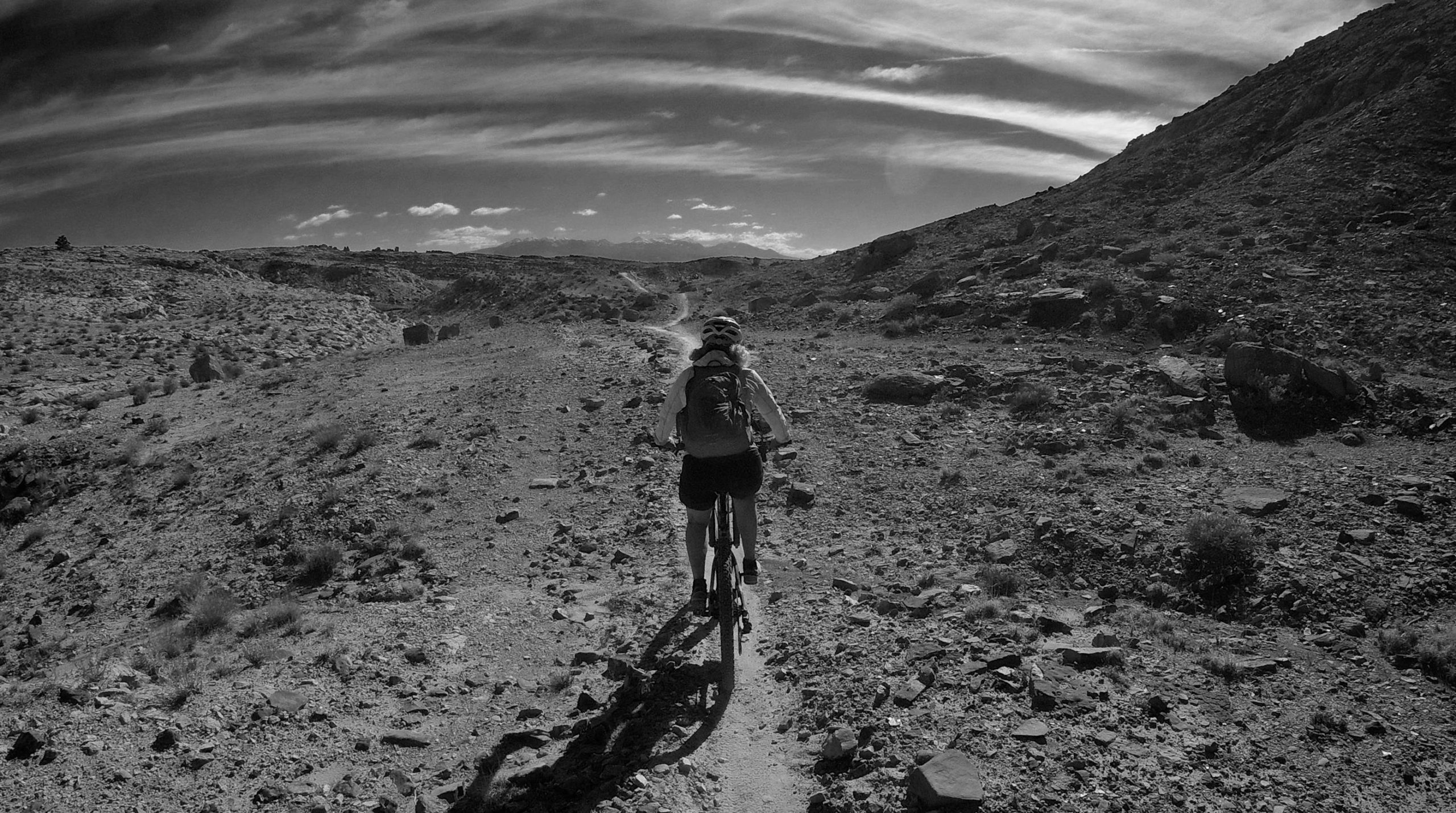 A cyclist riding a mountain bike on a rocky trail through a rugged landscape, with expansive skies and hills in the background. The image is presented in black and white. Klondike Bluffs mountain bike trail.