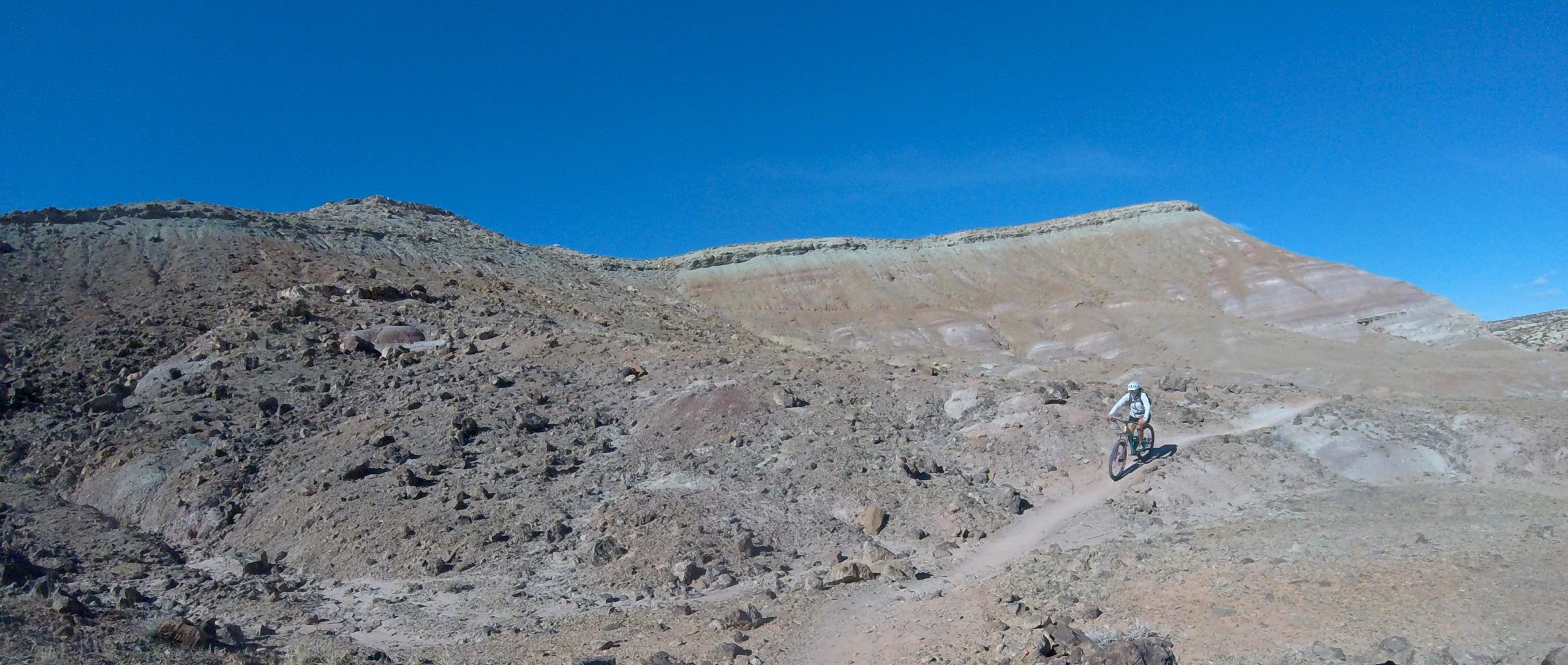 A mountain biker navigating a rocky trail in a barren landscape under a clear blue sky. The rugged terrain features varied rock formations and a distant hillside. Klondike Bluffs mountain bike trail.