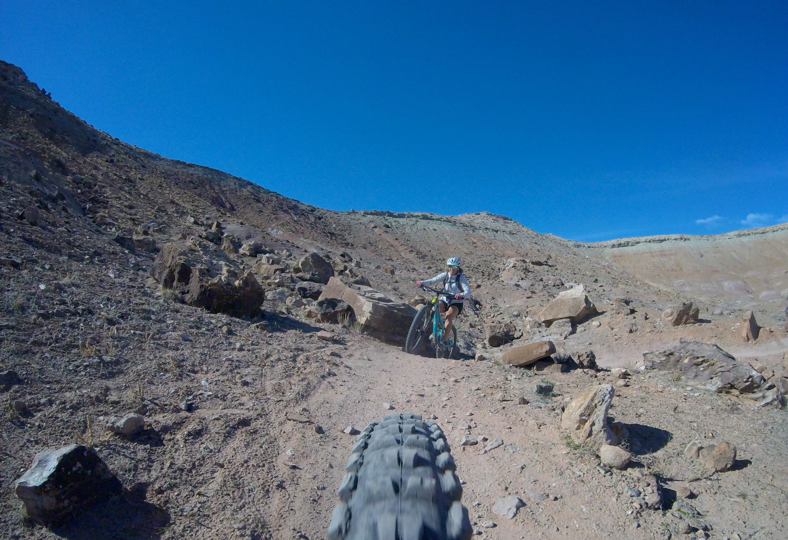 A mountain biker navigating a rocky trail in a desert landscape under a clear blue sky. The cyclist, wearing a helmet, is riding a teal bike with a visible tire in the foreground. The terrain features dry earth and scattered rocks. Klondike Bluffs mountain bike trail.