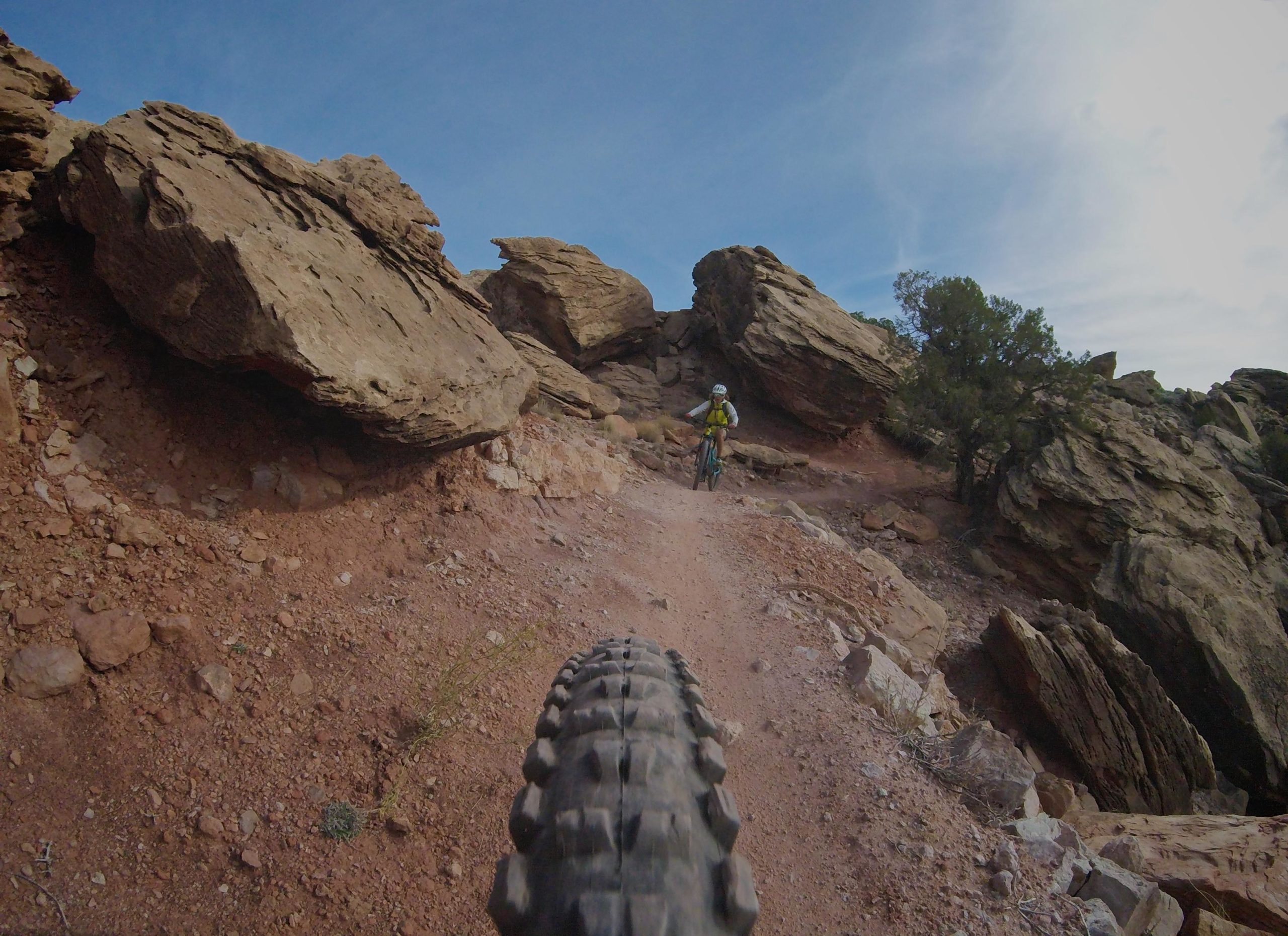 A mountain biker navigating a rocky trail with large boulders and red dirt beneath a blue sky. The image captures the bike tire in the foreground, emphasizing the rugged terrain. Klondike Bluffs mountain bike trail.