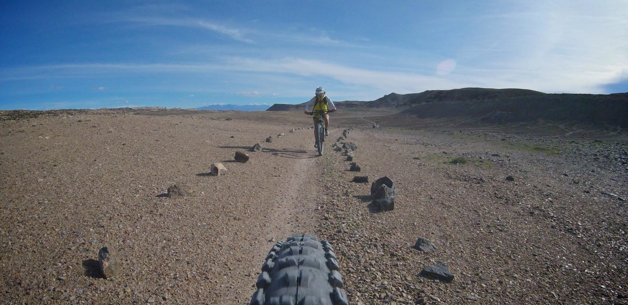 A cyclist riding on a gravel path through a desert landscape, with a clear blue sky overhead and mountains visible in the distance. The foreground features the tire of the bike and scattered rocks lining the trail. Klondike Bluffs mountain bike trail.