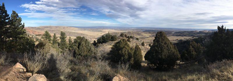 Panoramic view of rolling hills and valleys under a partly cloudy sky, surrounded by evergreen trees and brush. The landscape showcases a blend of natural vegetation and open terrain, creating a serene outdoor scene. Mount Falcon Park mountain bike trail.