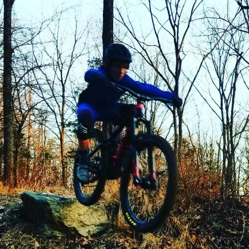 A young cyclist in a blue outfit jumps off a rocky surface while riding a mountain bike in a wooded area. Trees are visible in the background, with some foliage indicating a late fall or early spring setting. Nassau Trails mountain bike trail.
