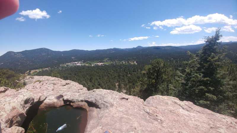 A panoramic view of a mountainous landscape taken from a rocky ledge, featuring lush green trees and a small town visible in the valley below, under a clear blue sky with a few clouds. A pool of water is visible in the foreground on the rock surface. 3 Sisters / Alderfer mountain bike trail.