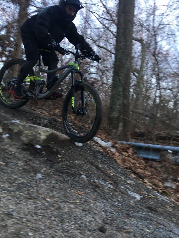 A mountain biker in a black outfit is navigating a rocky trail, mid-jump, with trees in the background. The rider is focused and airborne above a large rock, showcasing a dynamic action shot in a forested setting during winter. Meadowlark Park mountain bike trail.