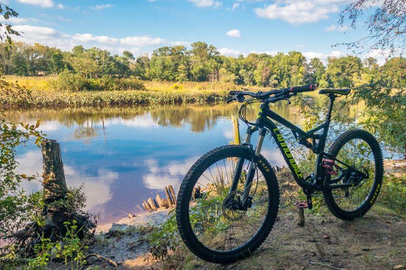 A mountain bike stands next to a calm river, surrounded by lush greenery and trees on a sunny day with a blue sky and fluffy clouds. The bike is positioned on a dirt path with a wooden stump and some plants in the foreground. Rancocas State Park - Westampton mountain bike trail.