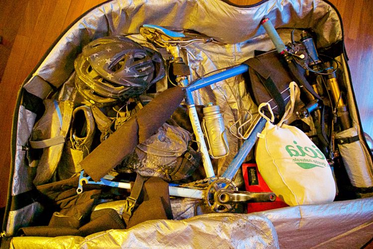 A packed bicycle travel case containing a blue bike frame with various components, a helmet, cycling shoes, a water bottle, and other gear, all secured in protective padding.