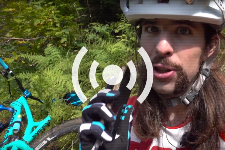 A person wearing a helmet and gloves is speaking while holding a small object, set against a backdrop of lush greenery and a blue mountain bike in the foreground. The image includes a play button graphic, suggesting it is a still from a video.