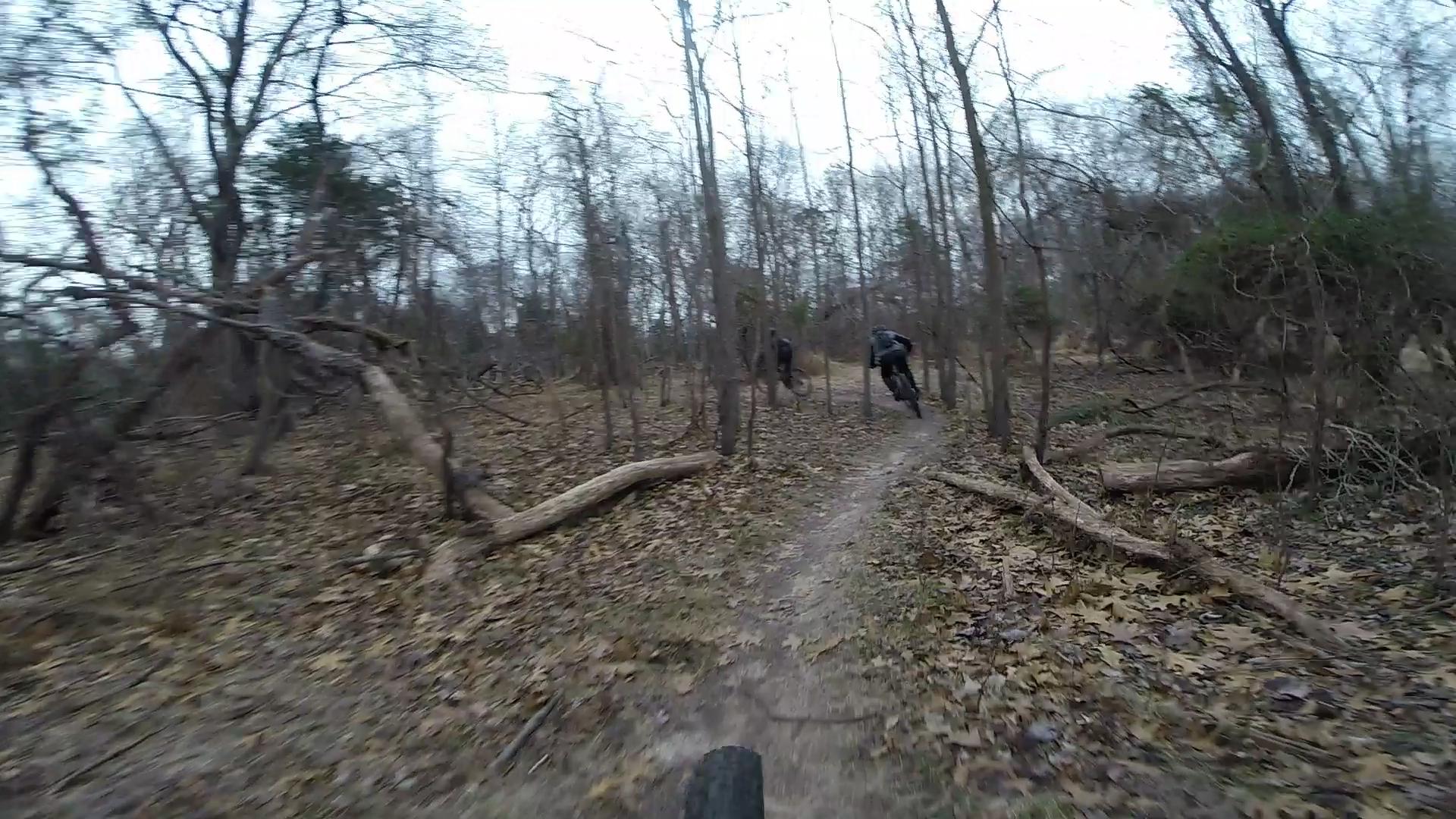 A mountain bike trail winding through a wooded area with bare trees and fallen leaves on the ground. Two cyclists are riding along the trail, navigating the path ahead. The scene conveys a sense of adventure and the beauty of nature in an autumn or winter setting. Allaire State Park mountain bike trail.