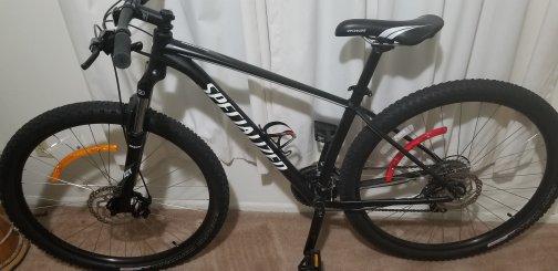 Specialized Rockhopper: A black mountain bike with disc brakes, featuring a sturdy frame and wide tires. The bike is positioned next to a window with white curtains, showcasing a sleek design with yellow and red reflectors on the wheels.