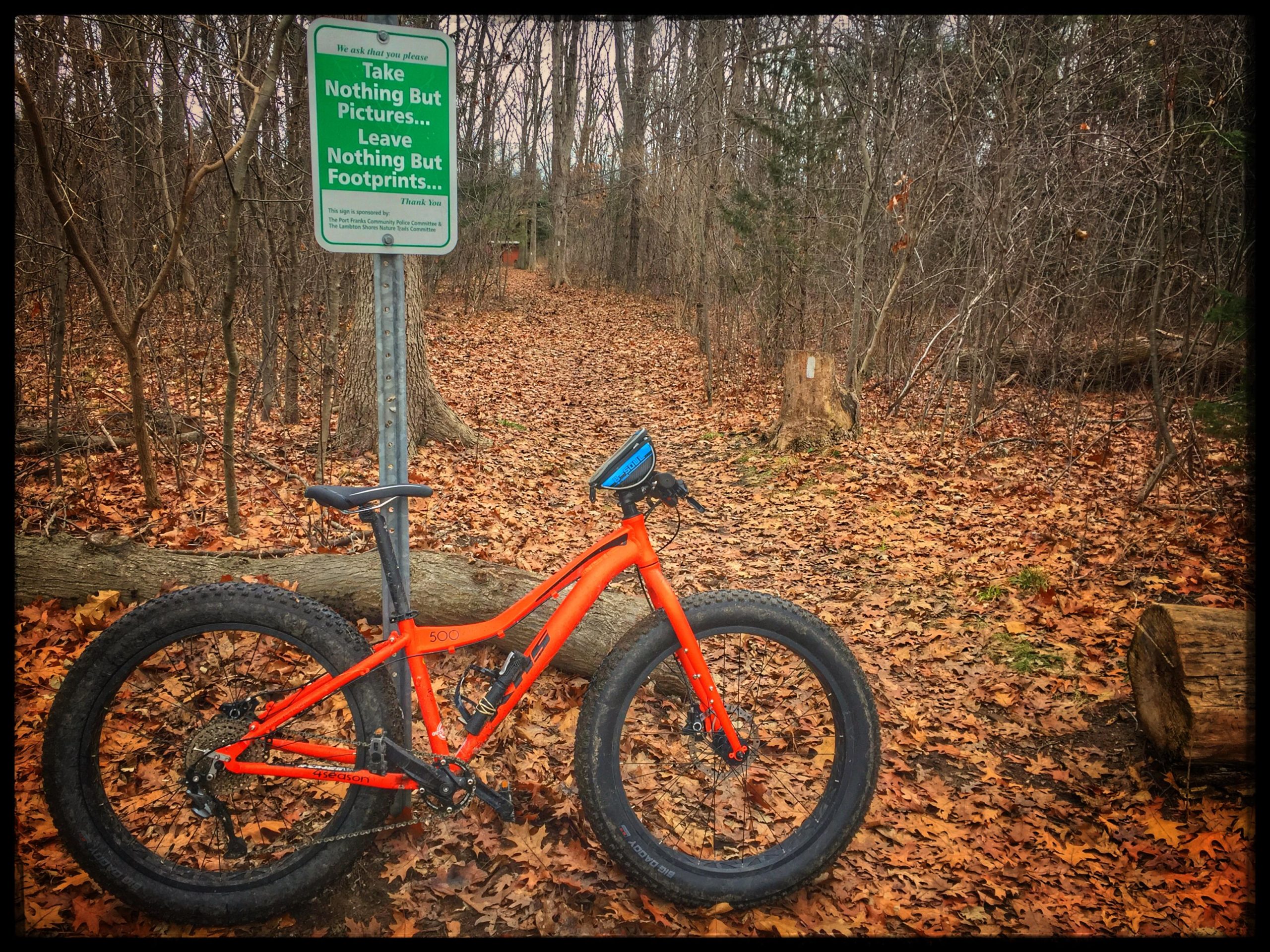 A vibrant orange fat bike rests against a wooden post in a forested area covered with brown leaves. In the background, a sign reads, “Take Nothing But Pictures… Leave Nothing But Footprints…” indicating a nature conservation message. The scene captures a tranquil outdoor environment with bare trees and fallen branches, suggesting a late autumn or early winter setting. Lambton County Heritage Forest mountain bike trail.