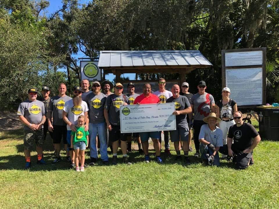 A group of individuals, including children and adults, stands together outdoors in front of a wooden shelter. They are wearing matching gray shirts with the "BMBA" logo. The group is holding a large novelty check made out to the City of Palmetto Bay for $41,250. The background features trees and information boards.