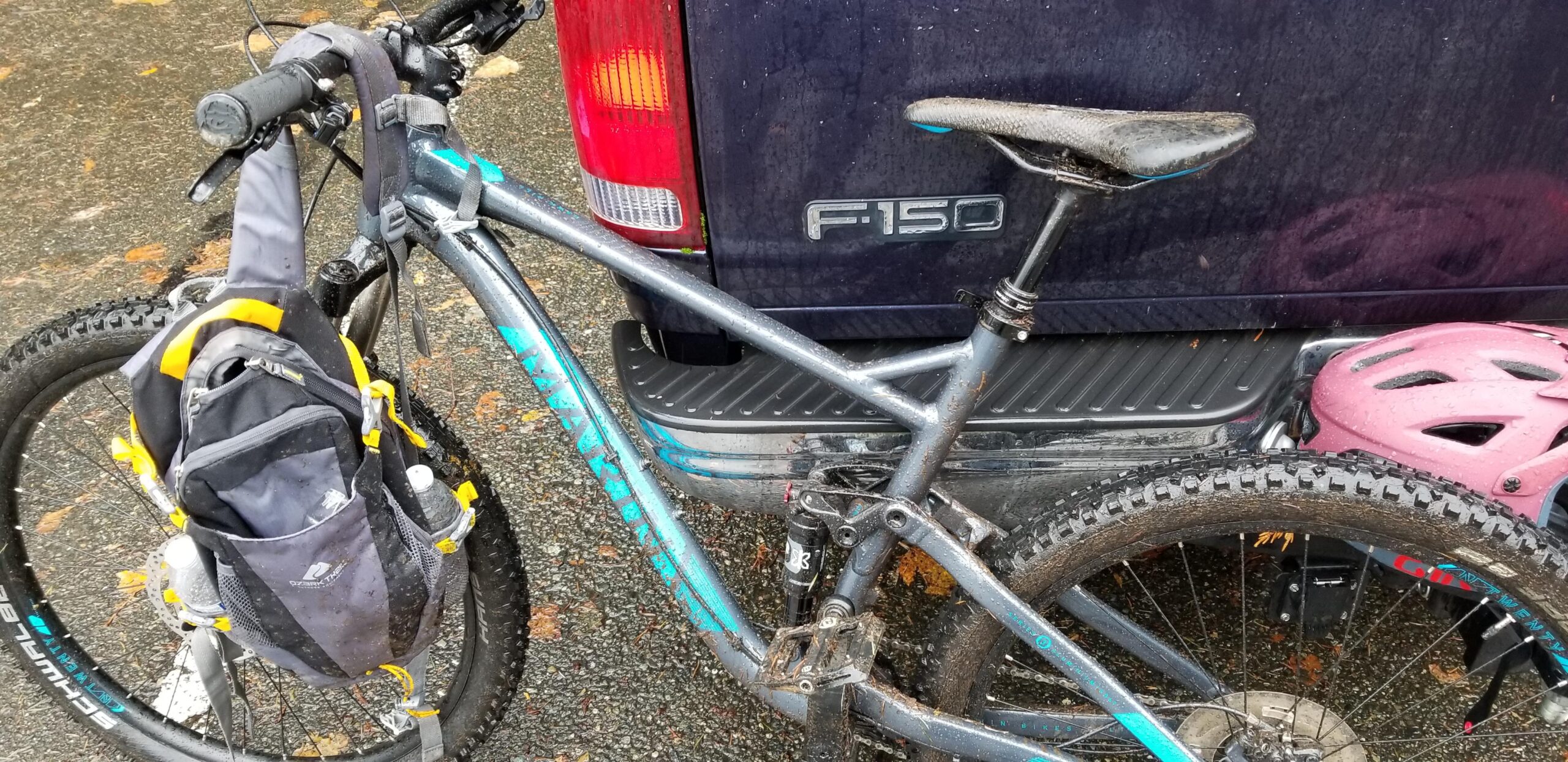 Marin Hawk Hill: A close-up view of a mountain bike parked next to a truck. The bike features a silver frame with blue accents and a black and yellow backpack attached to the seat post. A pink helmet is visible in the truck bed, and the scene appears to be wet, indicating recent rain. The ground is covered in small leaves.