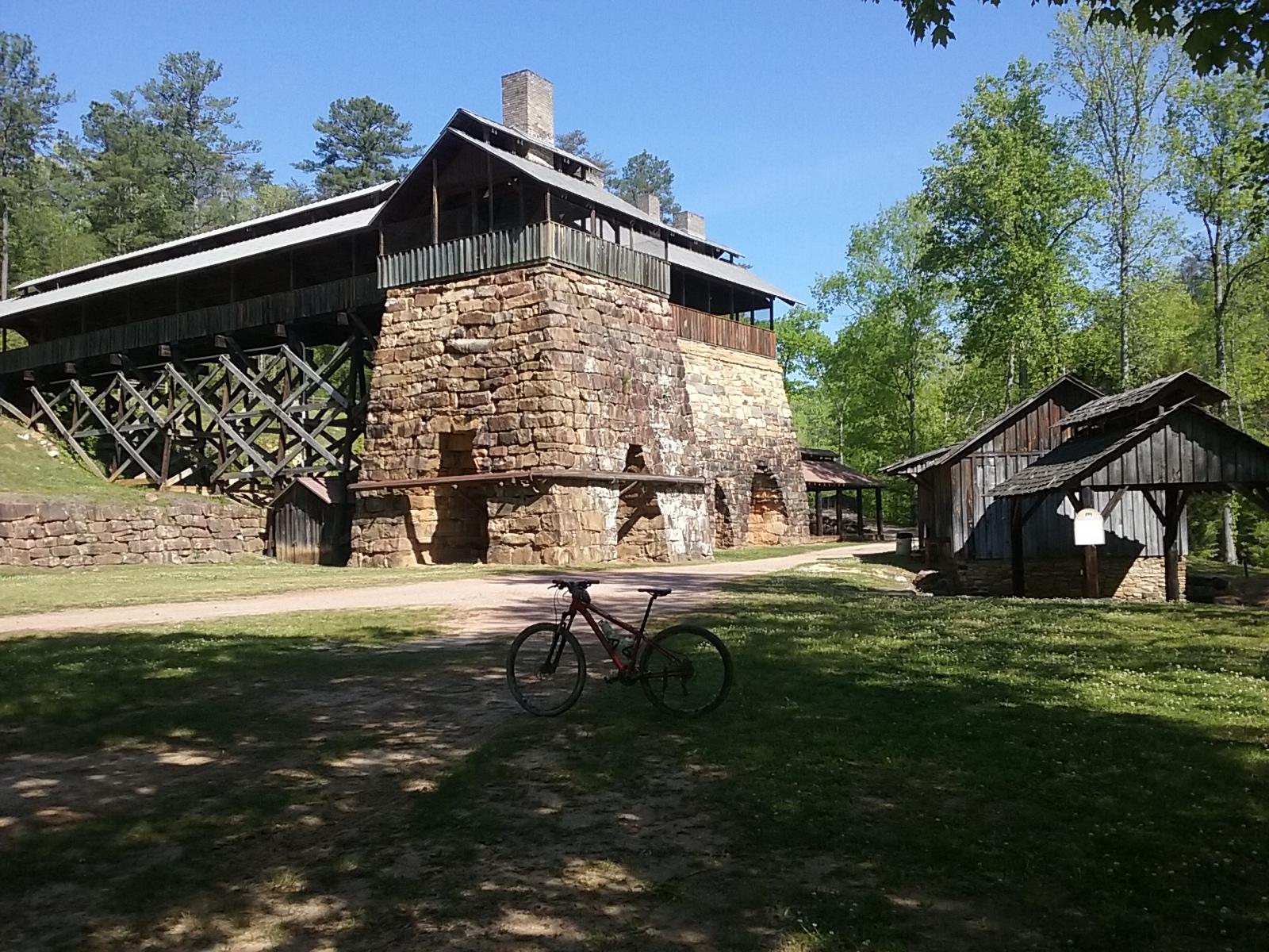 A historical structure with a stone base and wooden upper level is surrounded by lush greenery. In the foreground, a bicycle is parked on a dirt path, with smaller wooden buildings visible to the right. The sky is clear and blue, indicating a bright day. Tannehill Historic Ironworks State Park mountain bike trail.