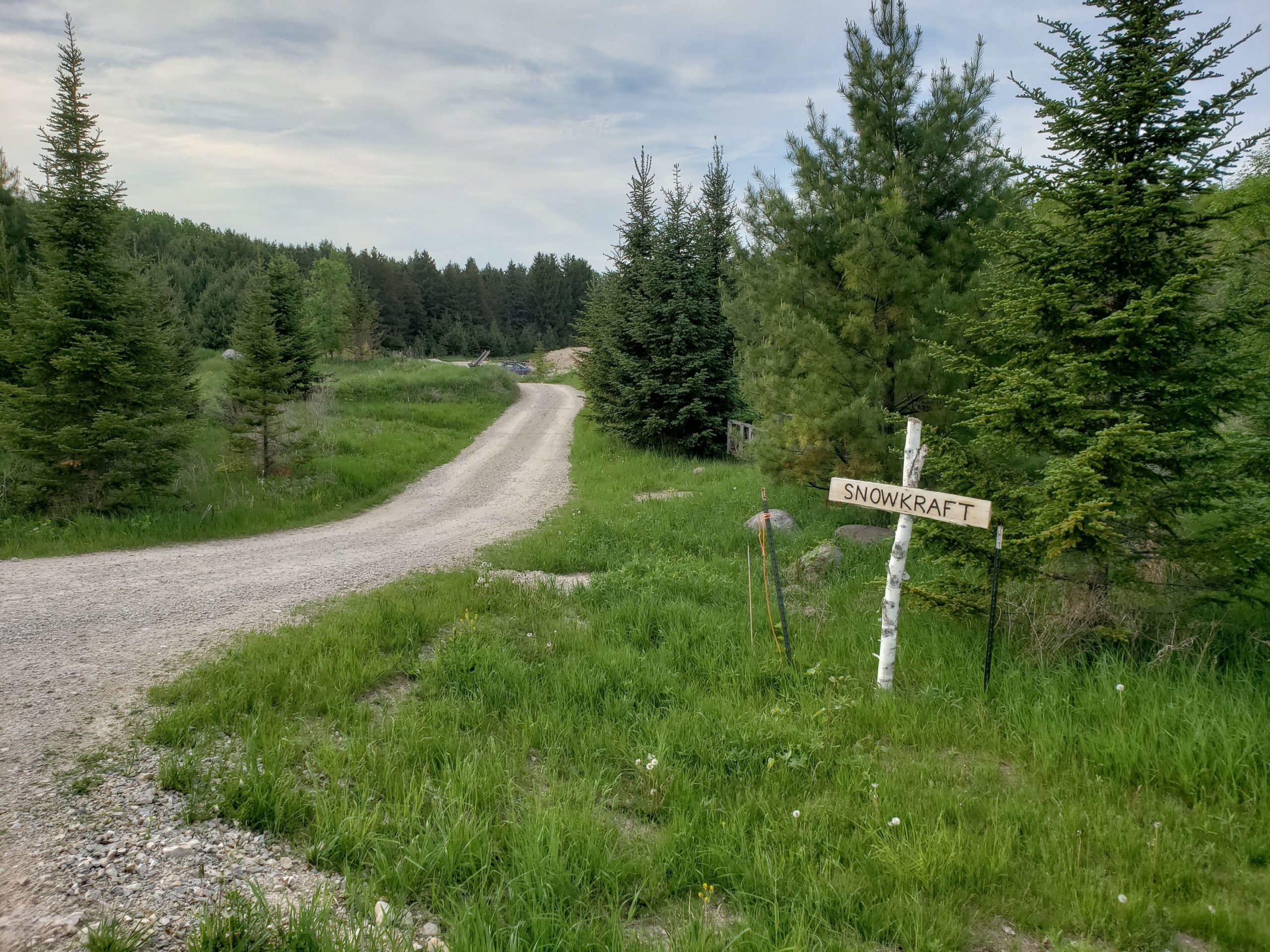 A dirt road winds through a green landscape, flanked by trees. A wooden sign reading "SNOWKRAFT" is planted in the grass on the right, pointing towards the road. [Private] Snowkraft Orchard Loop mountain bike trail.