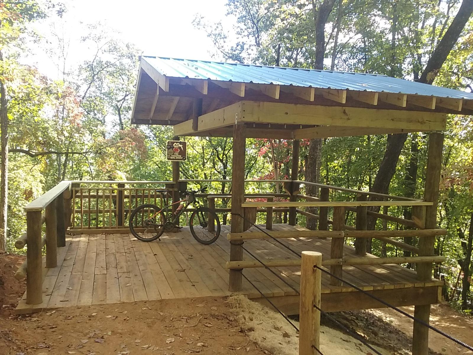 A wooden observation deck with a blue metal roof surrounded by trees, featuring a mountain bike resting against the railing. The deck offers a scenic view of the forest, showcasing autumn foliage. A sign indicating the area's purpose is visible. Fire Mountain Trail System mountain bike trail.