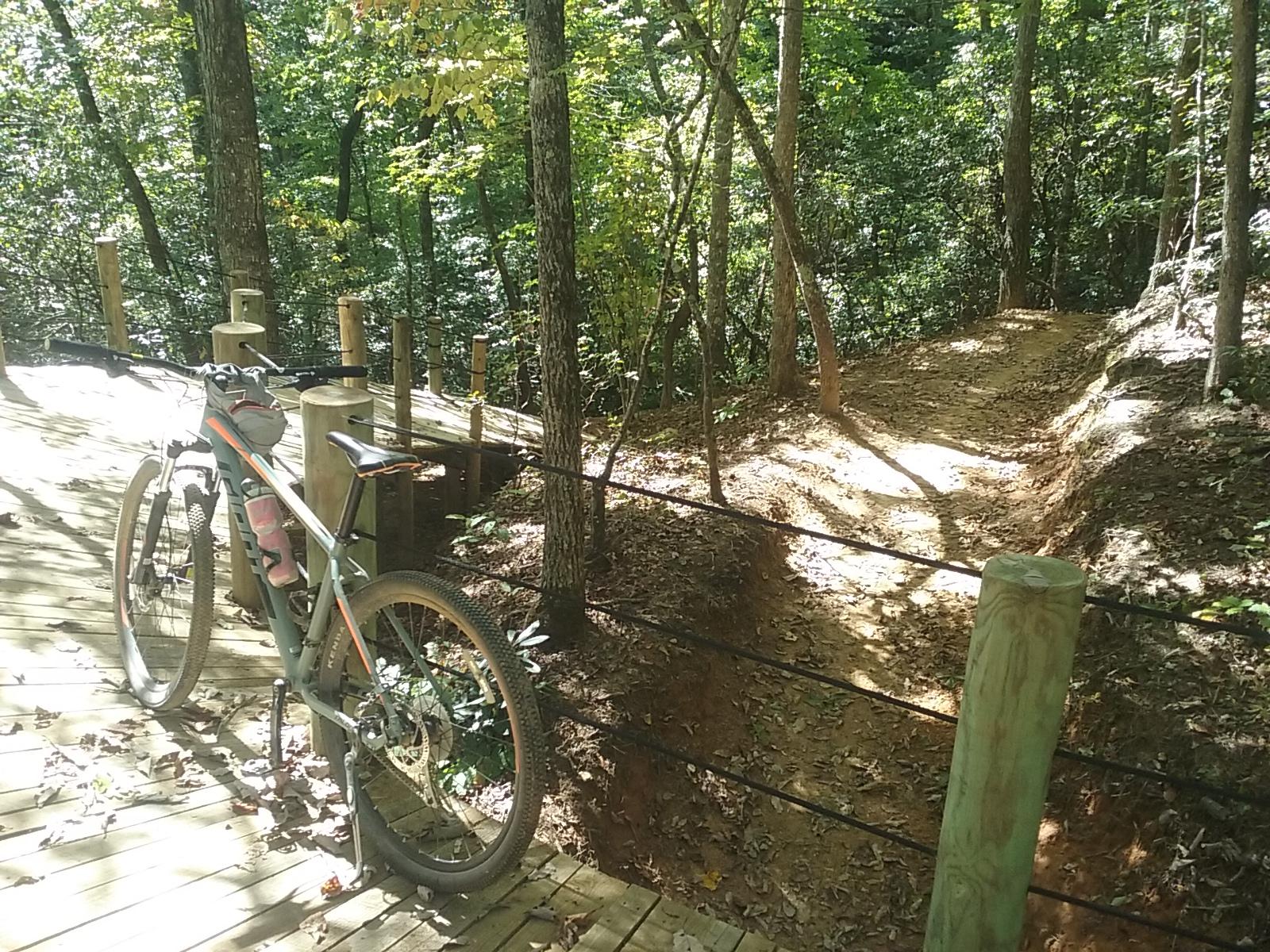 A mountain bike resting on a wooden deck trail in a forested area, with trees and foliage in the background. A dirt path diverges from the deck, leading deeper into the woods. Fire Mountain Trail System mountain bike trail.